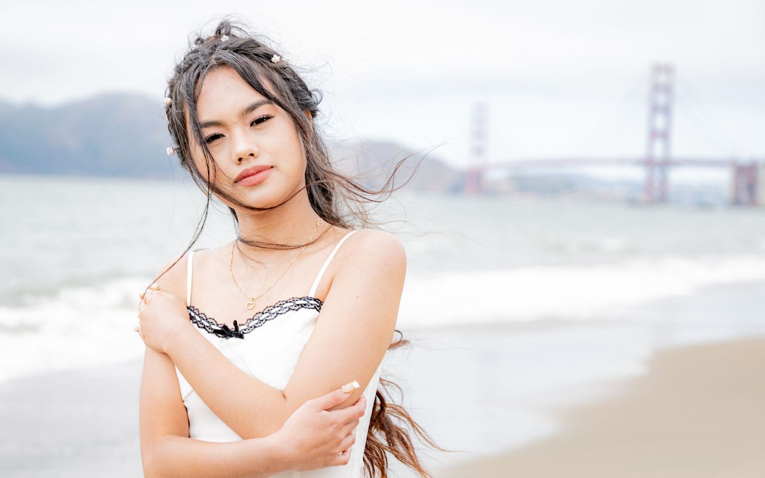 Young Asian woman in white dress crosses arms on beach with Golden Gate Bridge in soft focus behind.
