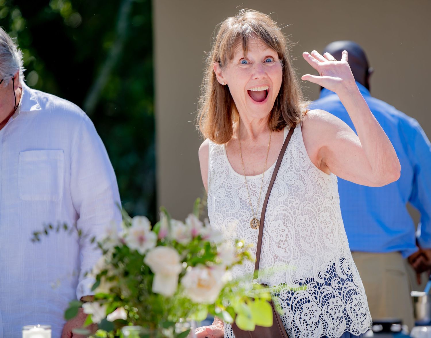 Excited woman in white lace top waving enthusiastically while holding floral bouquet outdoors.