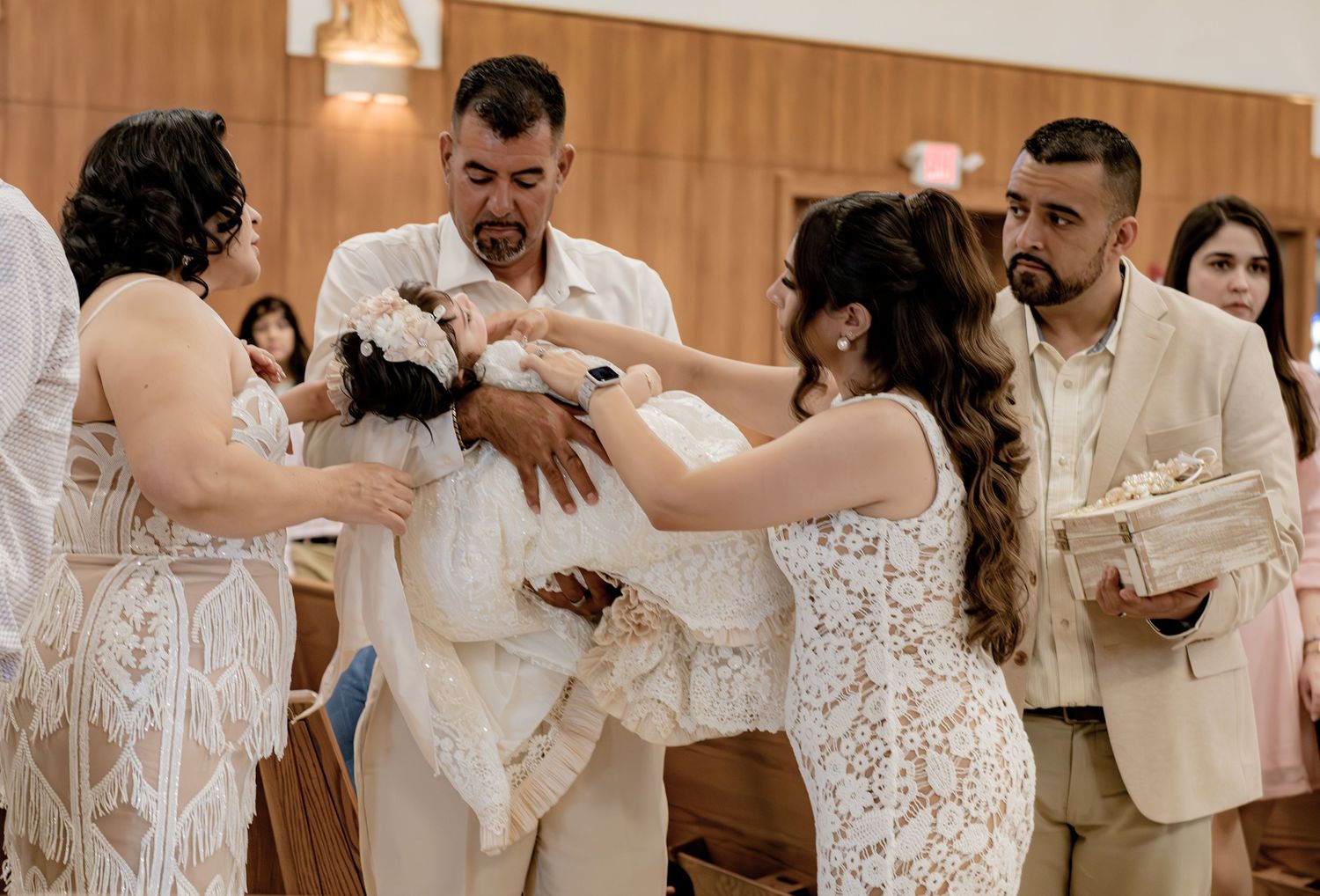Family and godparents gather around baby in white christening gown during a baptism ceremony in a Catholic church.