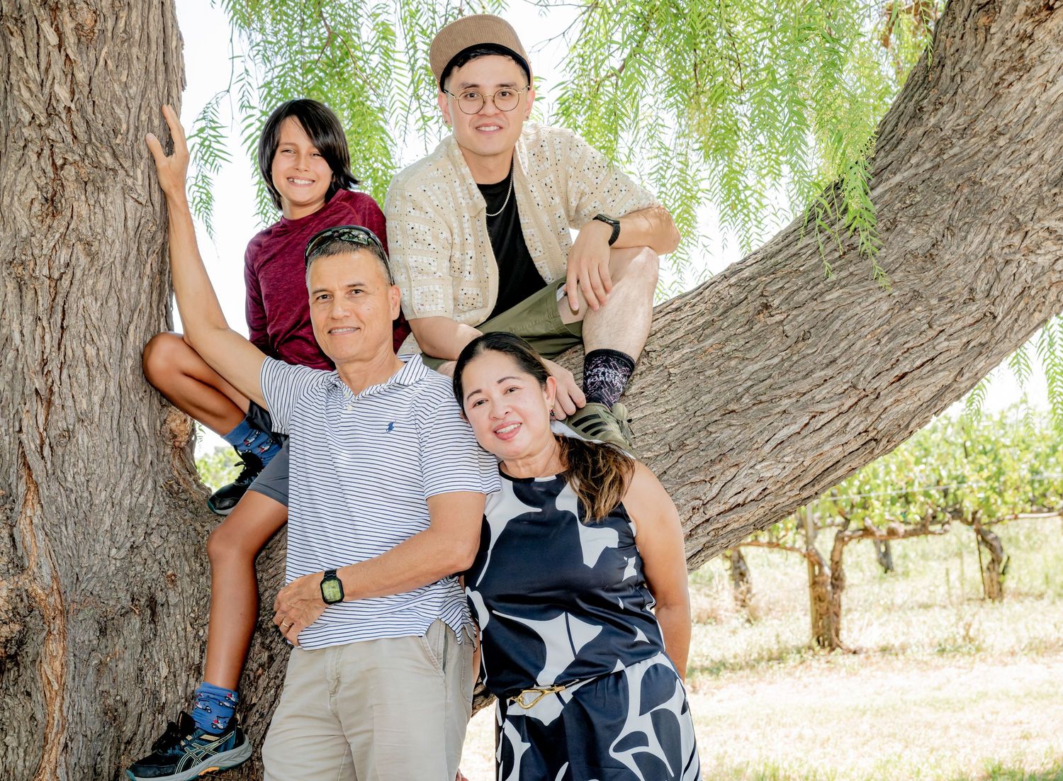 Family of four posing together on a large tree trunk outdoors in a sunny garden setting.