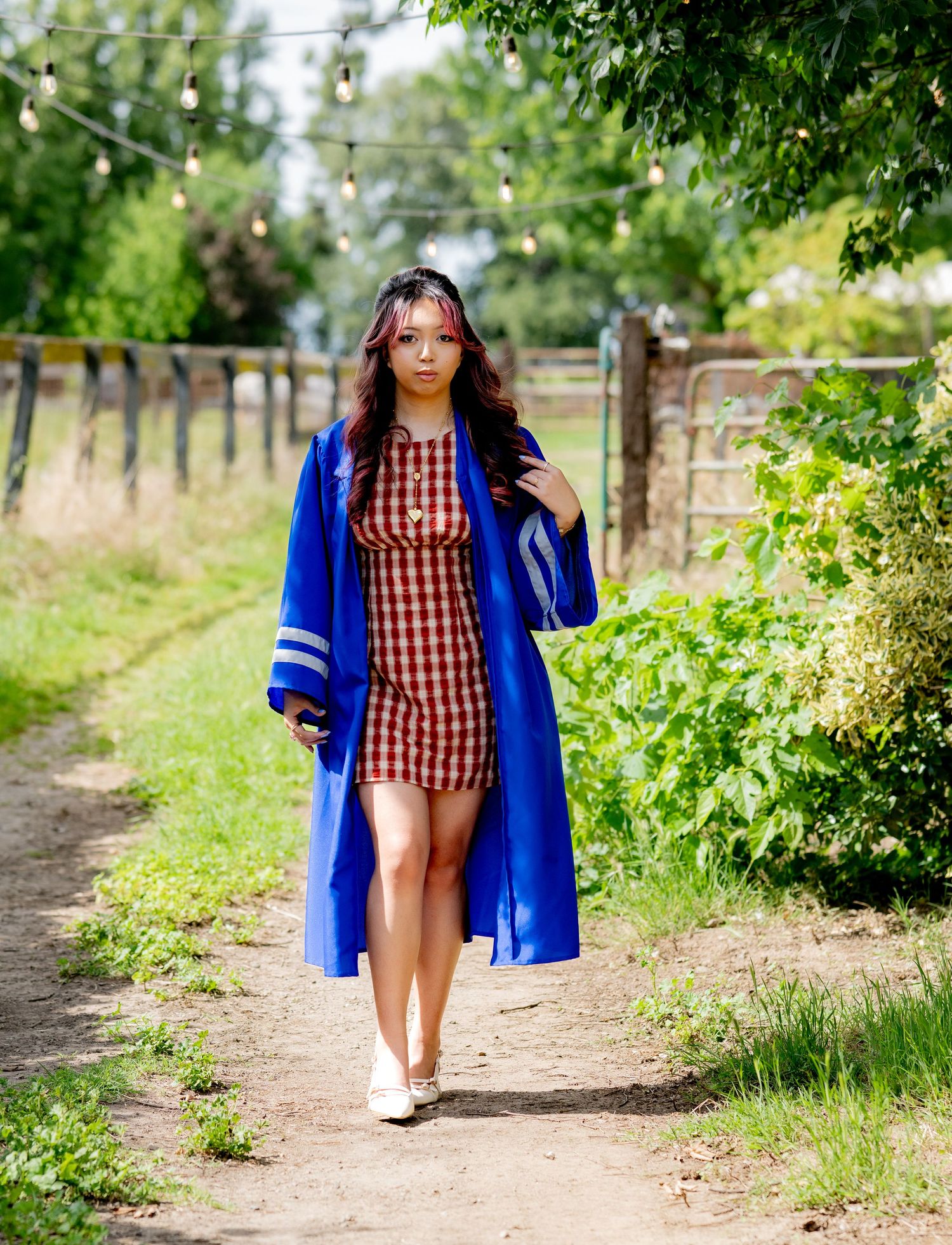 Graduate in blue commencement robe and red gingham dress walks along sunny garden path.