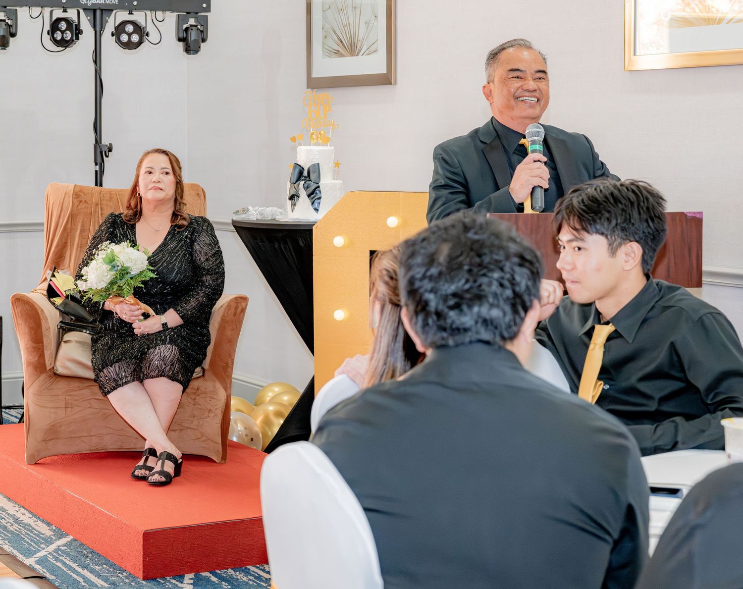 Man speaking with microphone at a 60th birthday celebration while woman sits holding flowers.