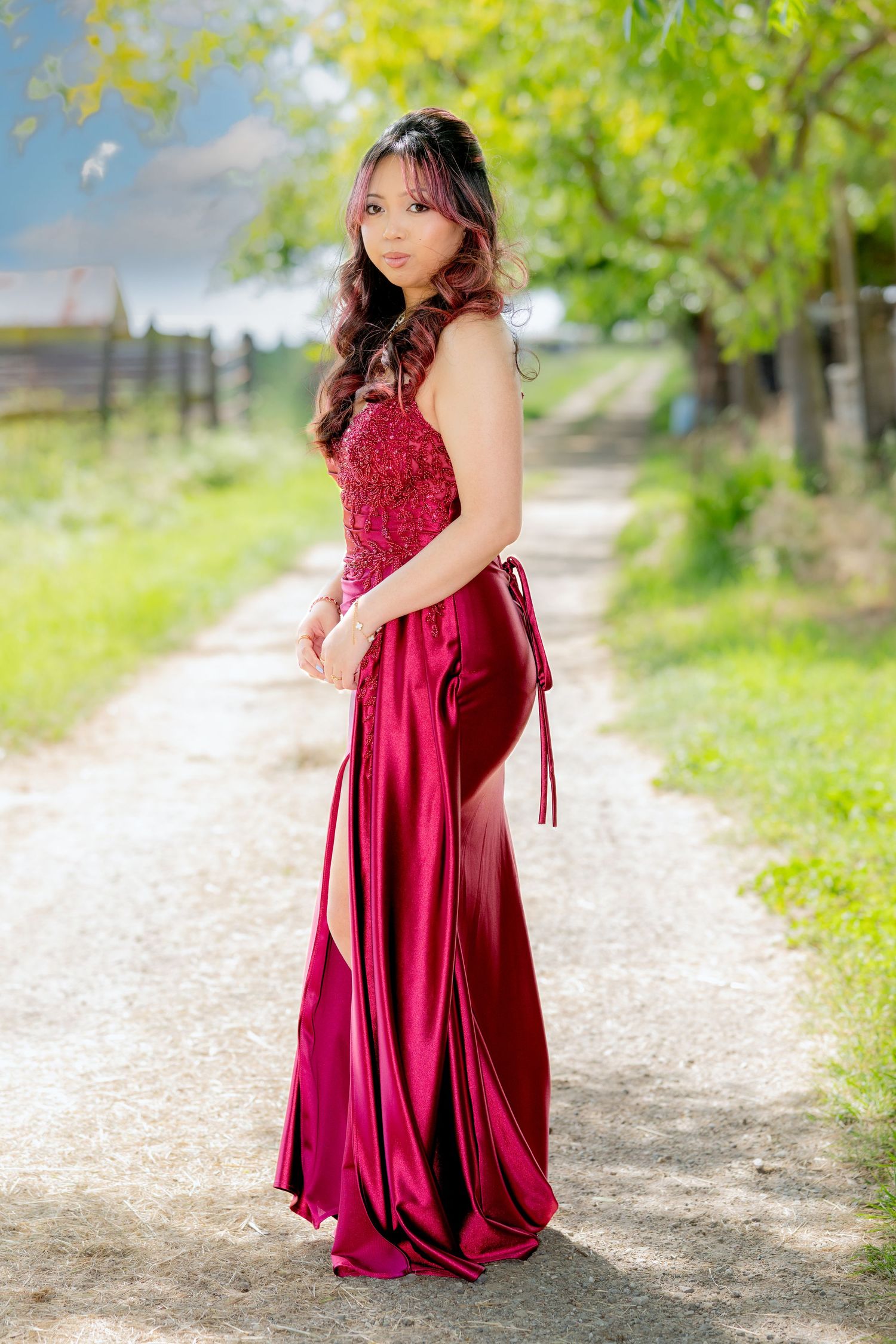 Asian woman in stunning crimson formal gown stands gracefully on a tree-lined path.