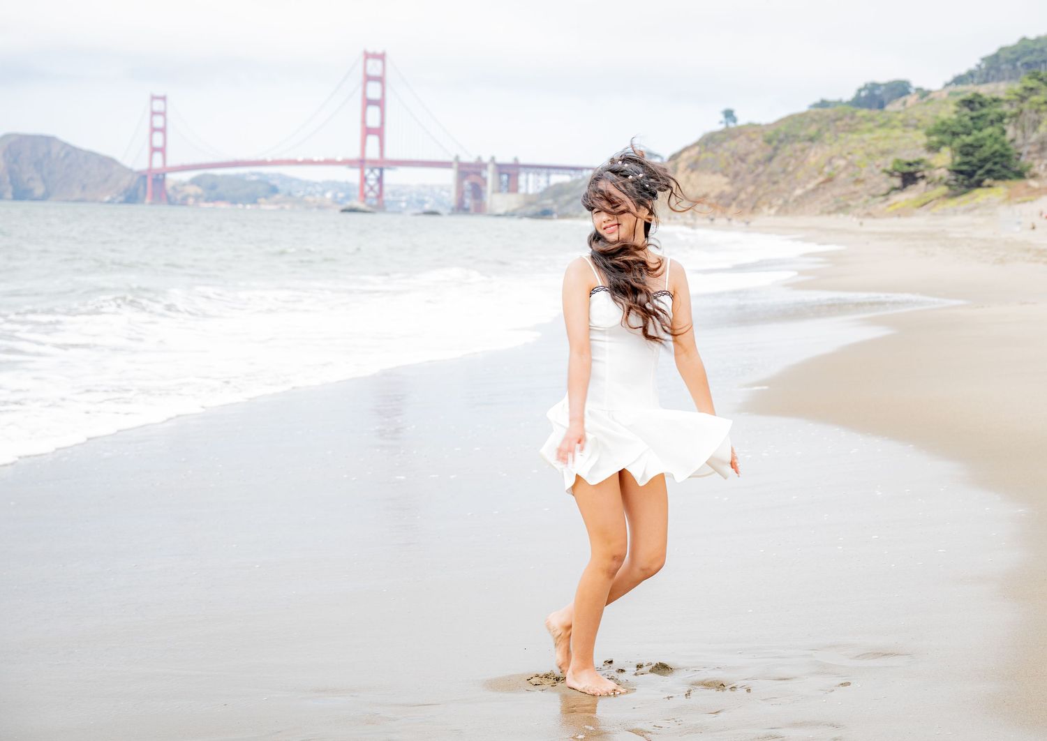 Woman in white dress walks barefoot on Baker Beach with Golden Gate Bridge visible behind her.