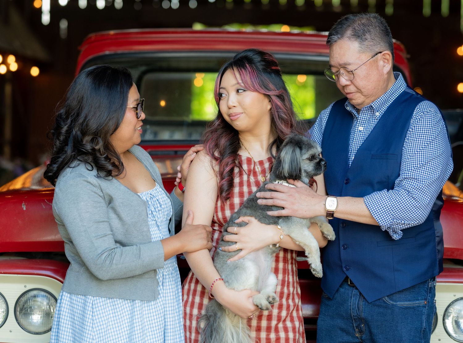Family interacting with small gray dog in front of vintage red truck in rustic barn setting.