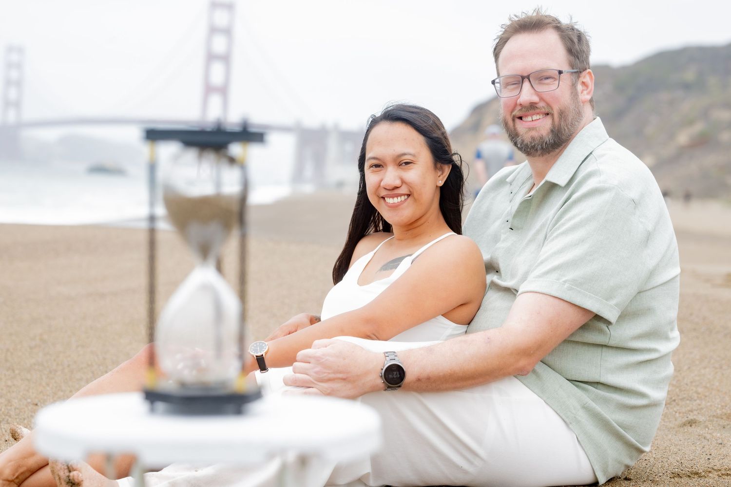 Smiling couple sitting on sandy beach with hourglass on white stool and Golden Gate Bridge in background.