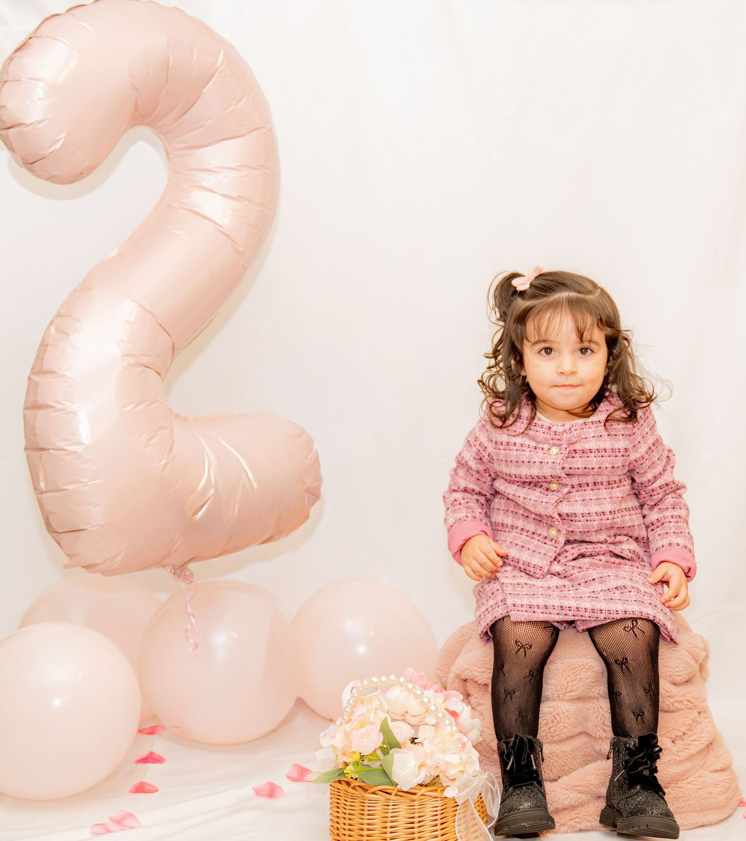 Toddler girl in pink tweed dress sits beside a number 2 balloon at her 2nd birthday party.