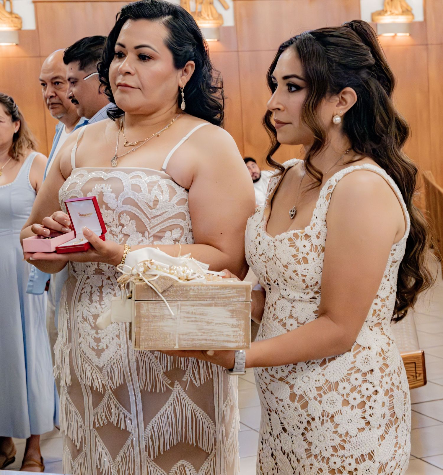 Two women in elegant dresses hold baptism gifts including a red jewelry box during a Catholic christening ceremony.
