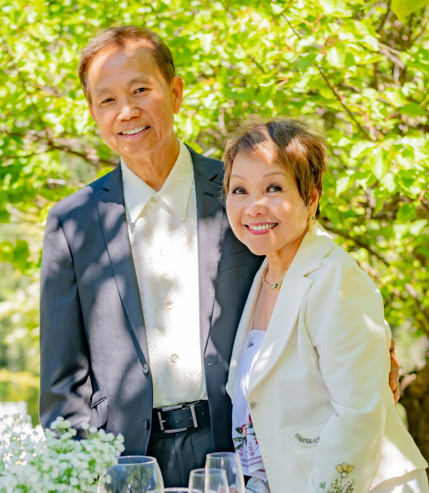 Smiling Asian couple posing together outdoors at a sunny garden event with wine glasses on table.