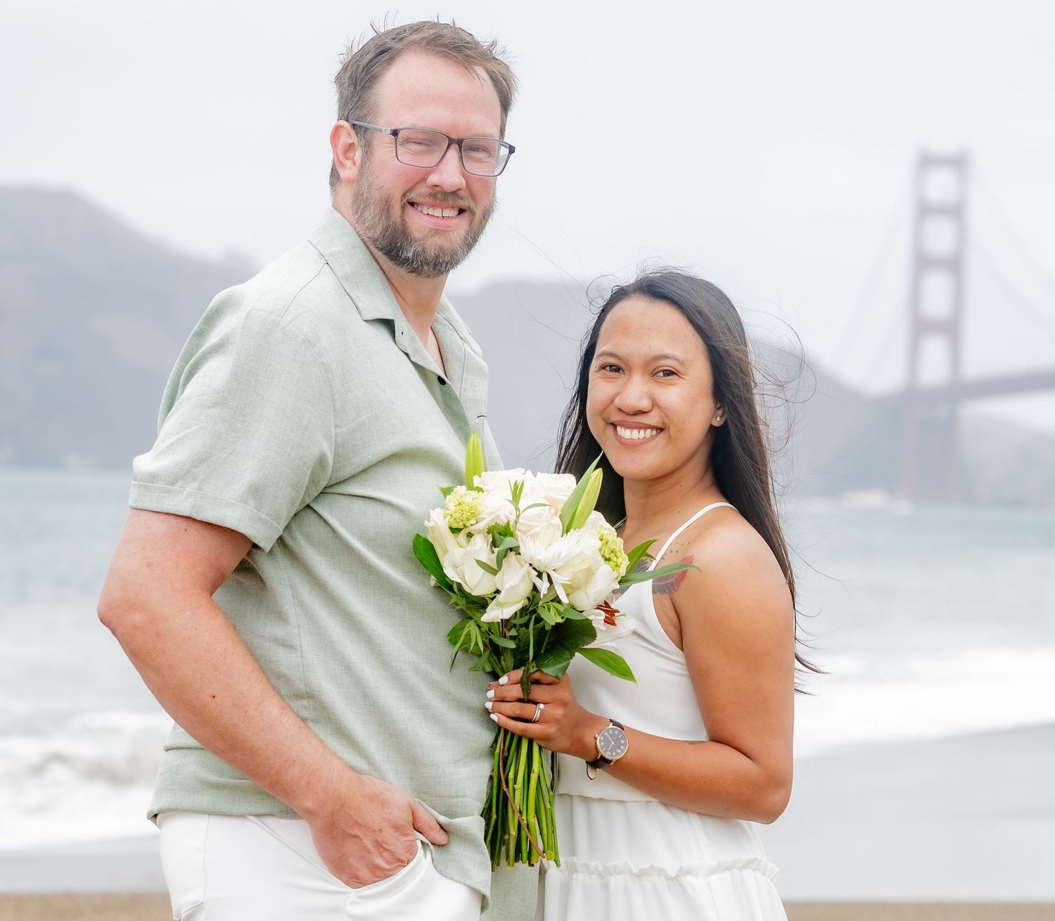 Happy couple poses with white floral bouquet on Baker Beach with Golden Gate Bridge behind them.