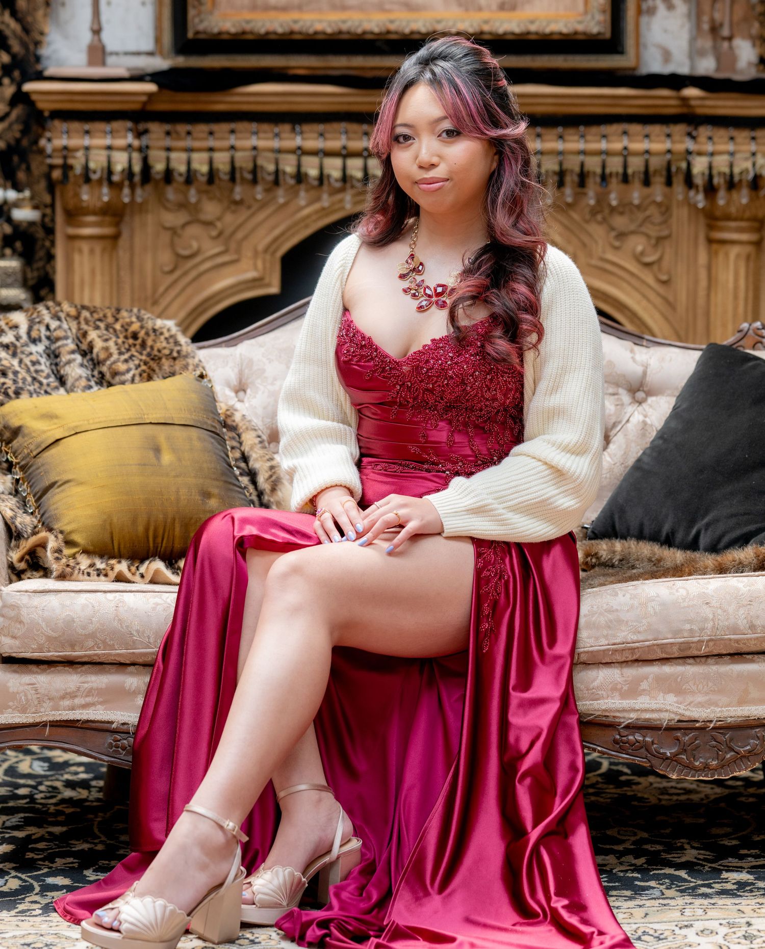 Young woman in crimson satin dress and cream cardigan sitting elegantly on a vintage chaise lounge.