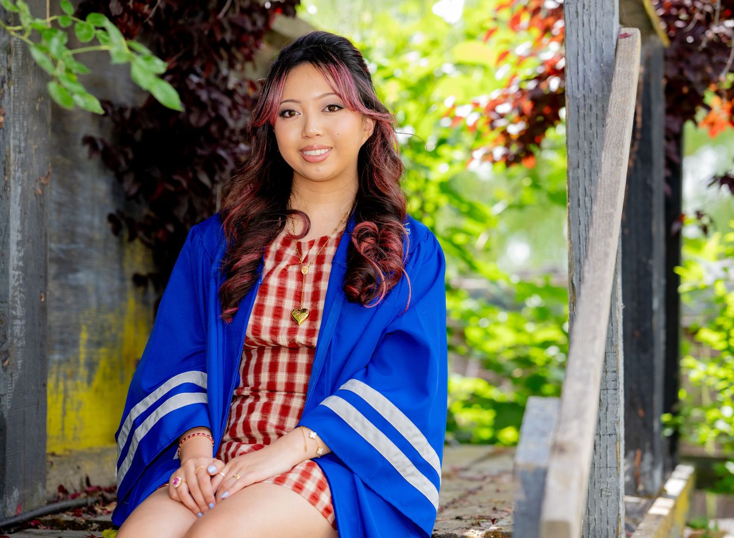 Young woman in blue graduation robe and red checkered dress seated in garden gazebo.