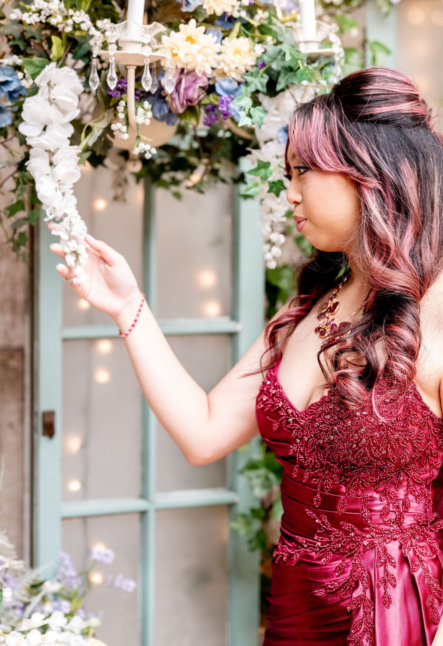 Young woman in burgundy beaded gown reaches for hanging white florals beneath chandelier in garden setting.