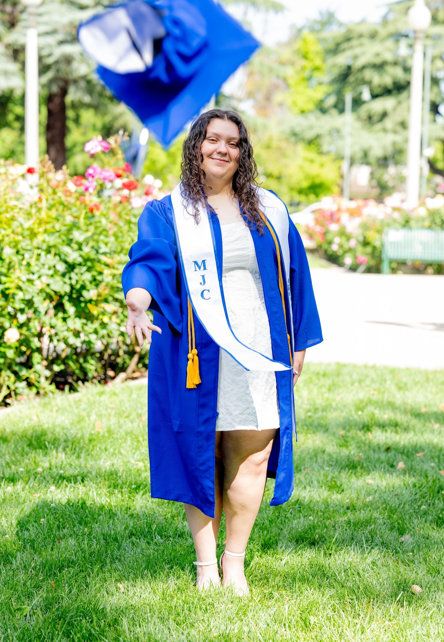 Smiling graduate in blue MJC cap and gown celebrates outdoors in a garden setting.