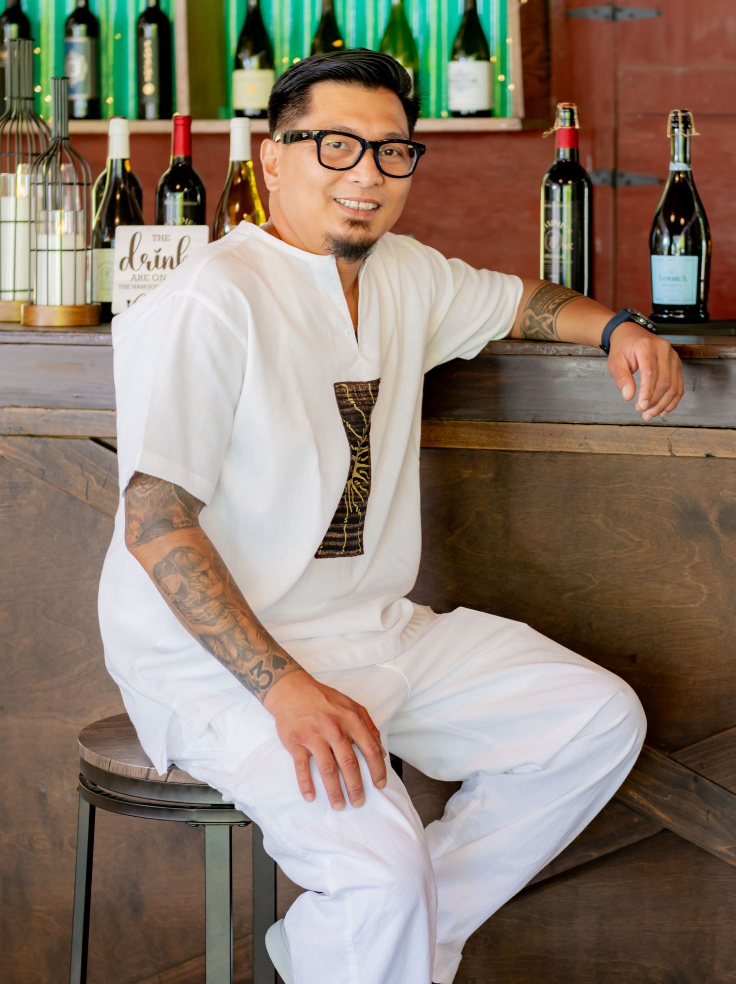 Tattooed man in white outfit sitting at a bar surrounded by wine bottles at an indoor venue.