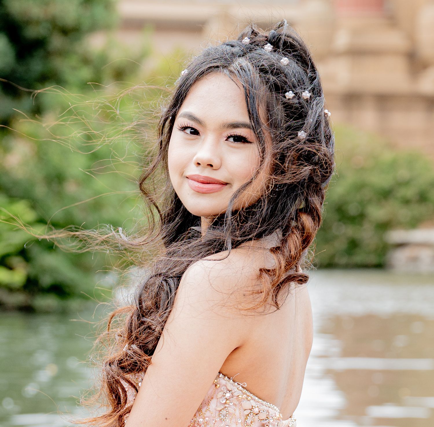 Young girl in blush strapless gown with floral hair accessories posing near waterfront.