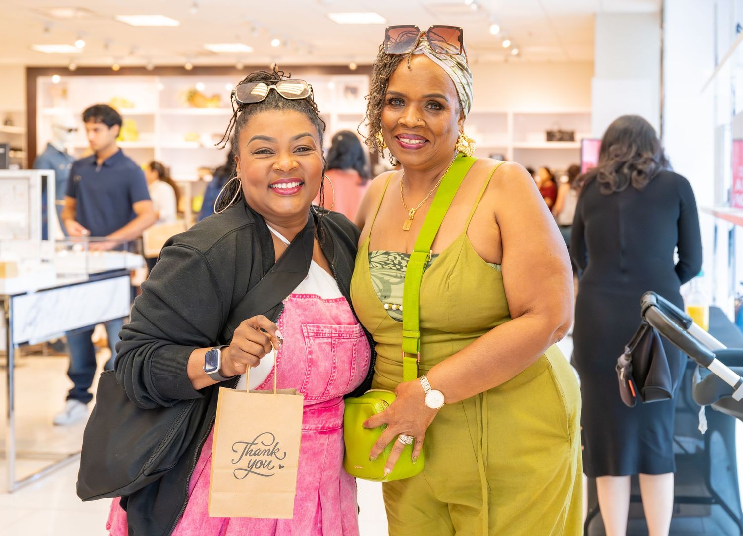 Two women smile together in retail store, one in pink dress holding thank you gift bag.