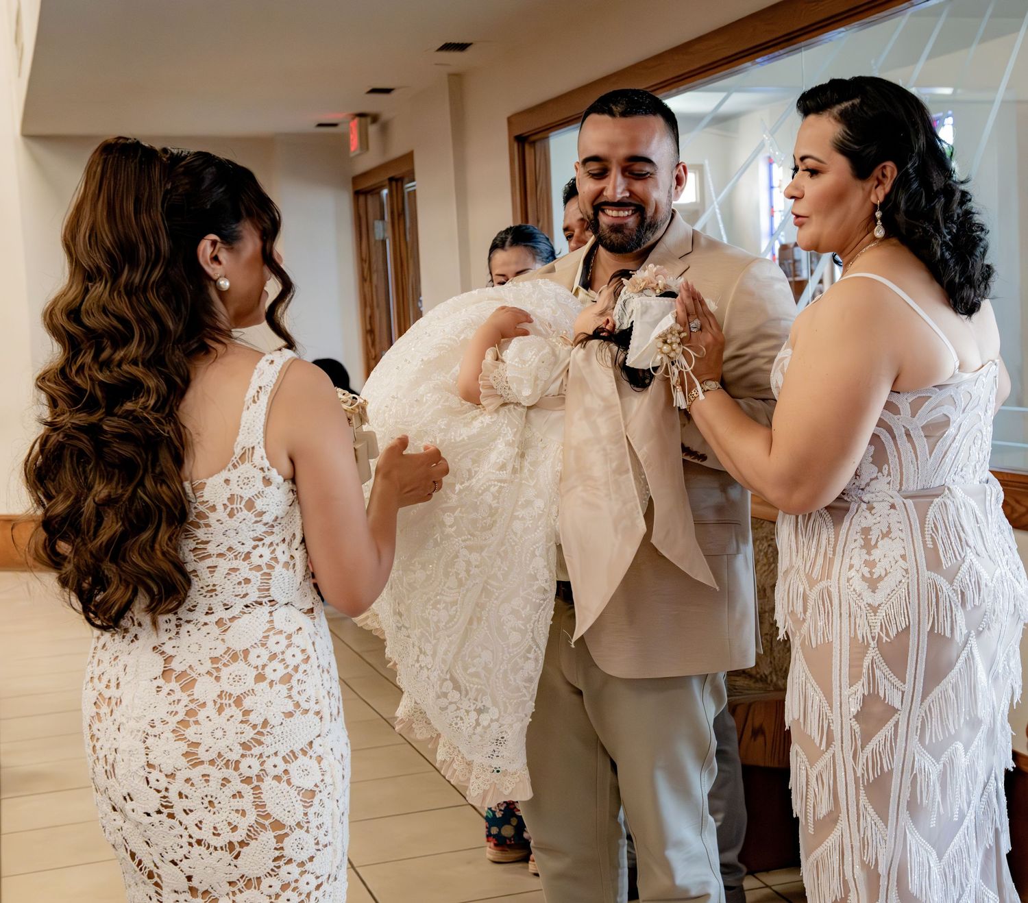 Family members joyfully admire baby in white lace christening gown during a baptism celebration in a church hallway.