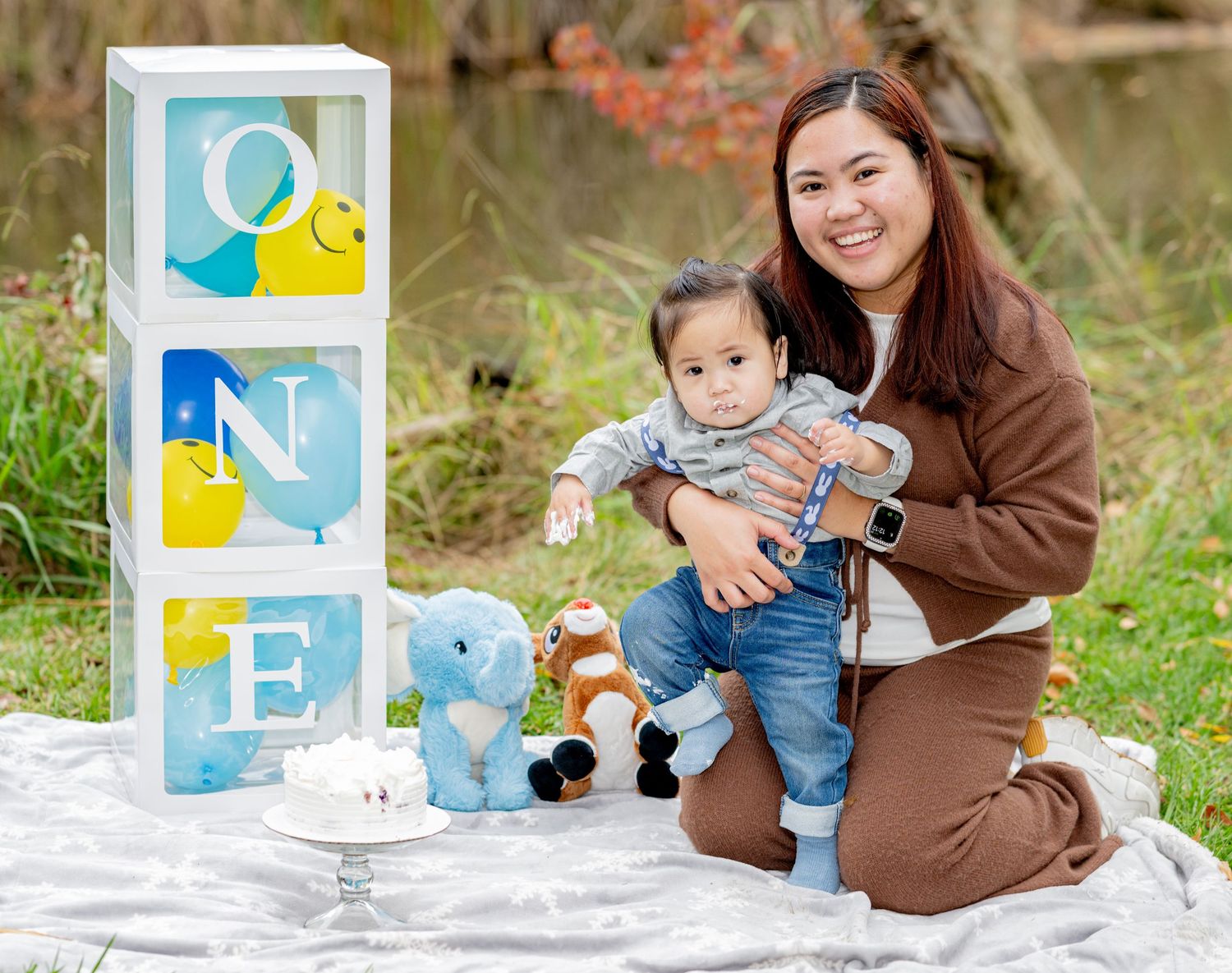 Smiling mom and baby boy posing at first birthday cake smash with ONE letter balloon boxes outdoors.
