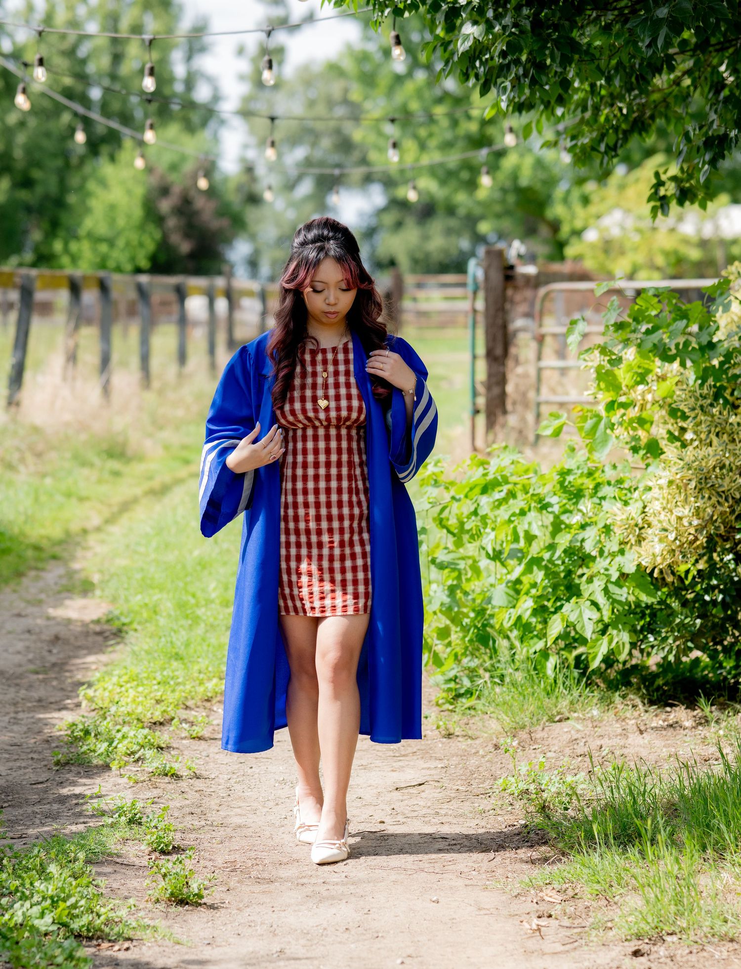 Graduate in blue gown over red plaid dress walks down garden path looking downward.