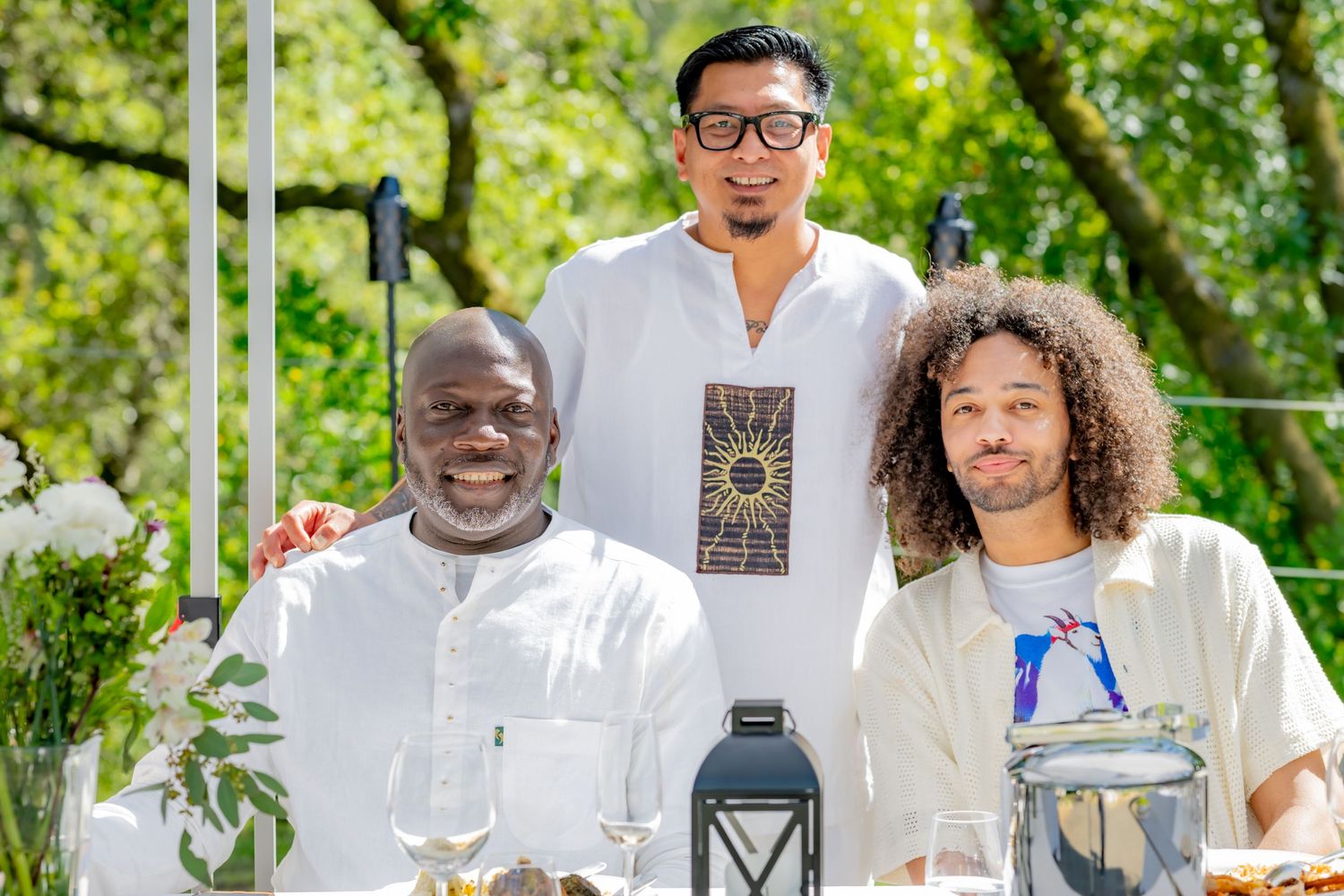Three men in white attire posing together at an outdoor dining event.