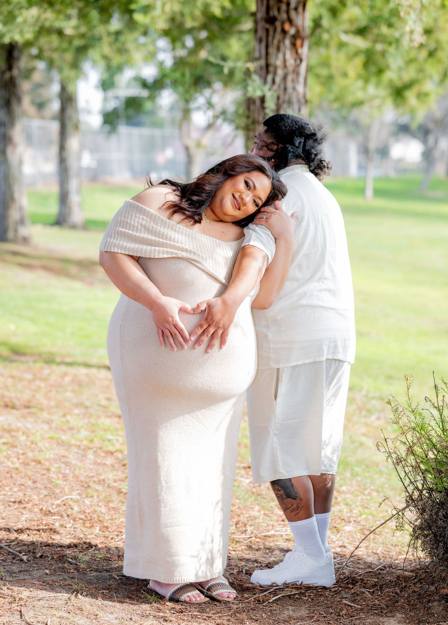 Man tenderly rests his head on pregnant partner's shoulder while she smiles, both in a sunlit park.