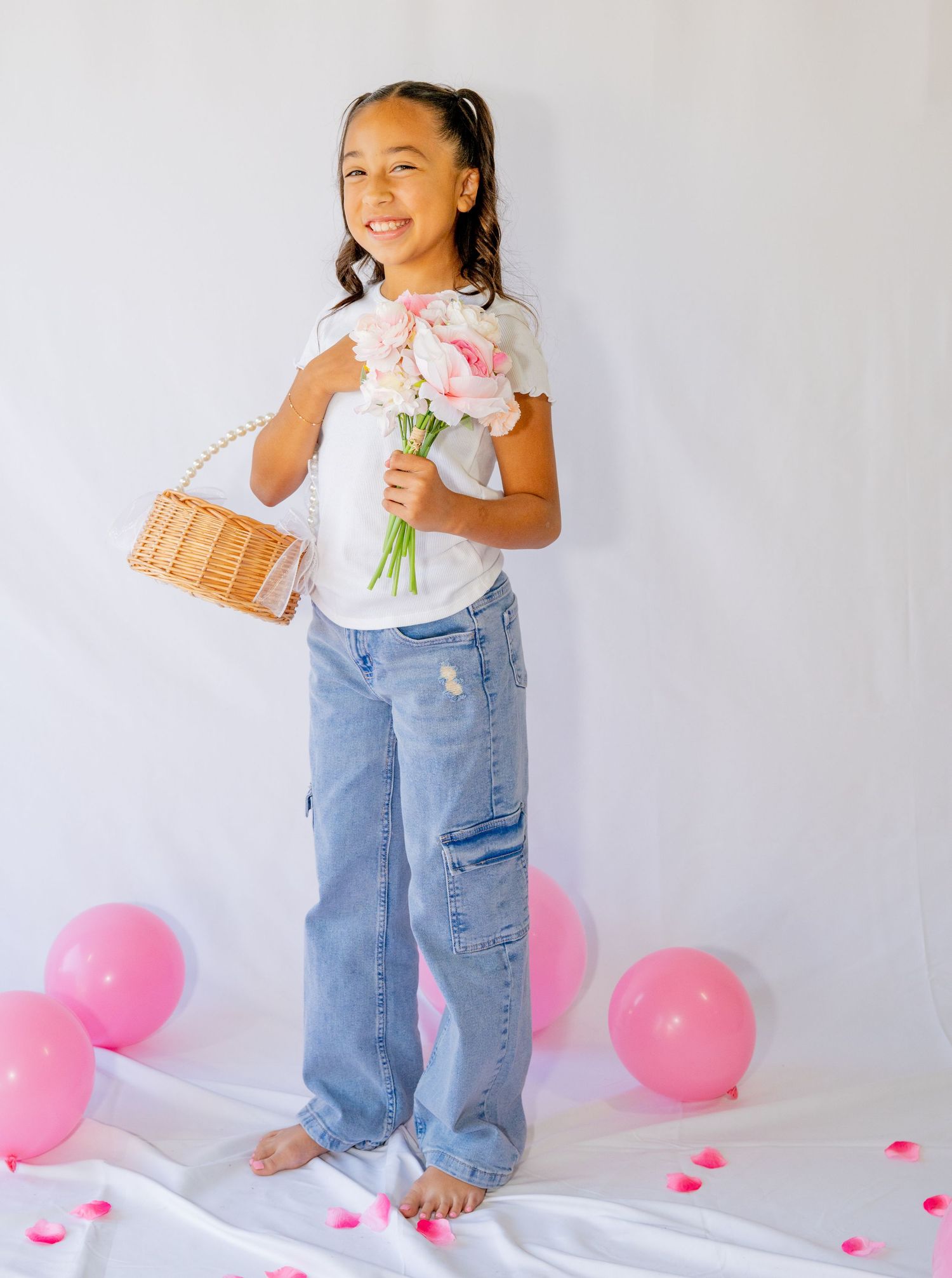 Young girl holding pink flowers and wicker bag surrounded by pink balloons on white backdrop.