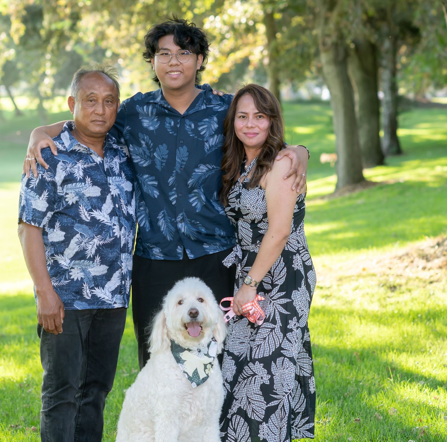 Family of three posing outdoors in a park with their white fluffy dog on a sunny day.