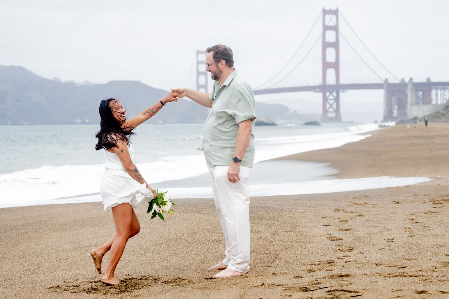 Smiling couple holding hands and dancing on Baker Beach with Golden Gate Bridge in the background.