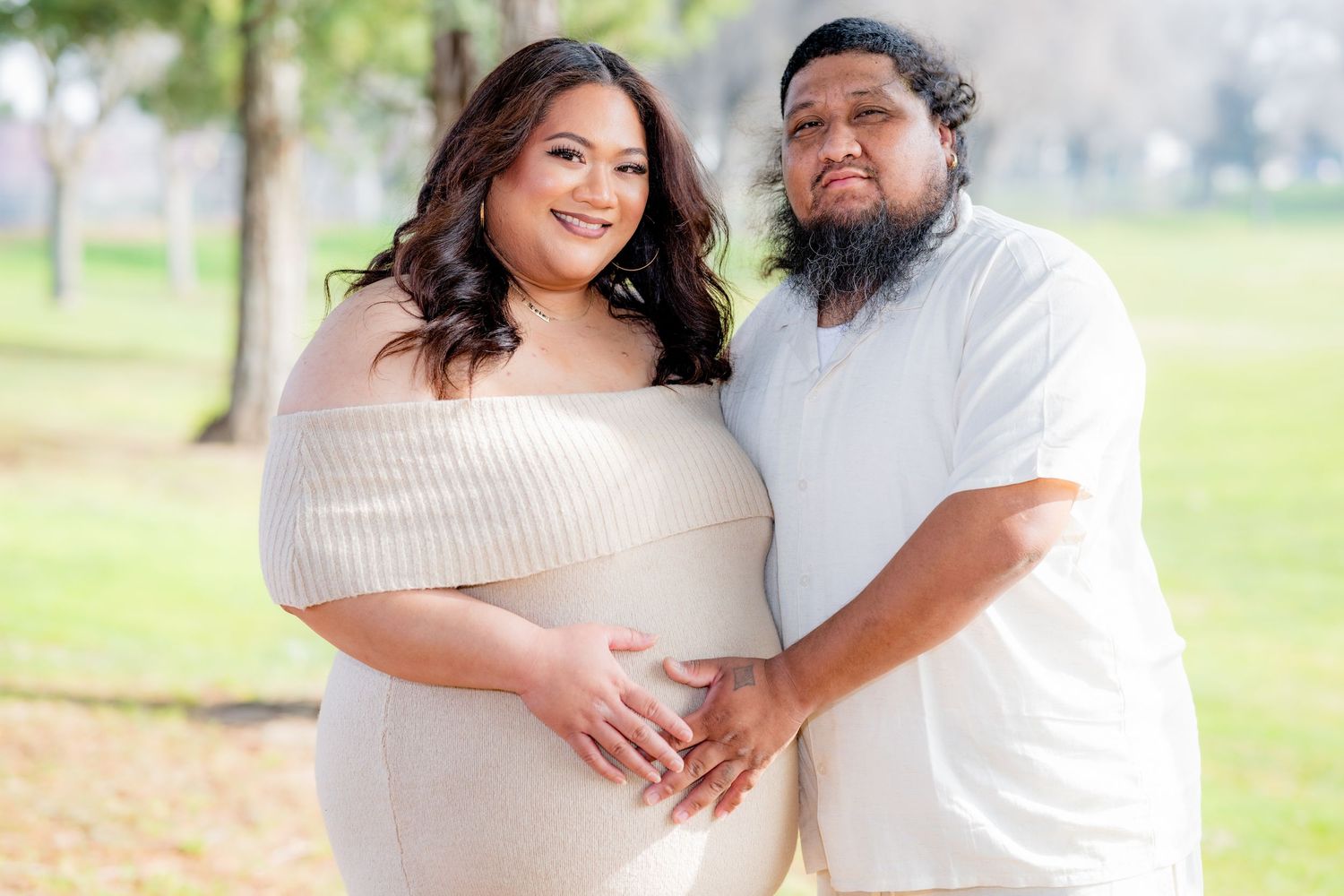 Smiling expectant couple in coordinating cream outfits cradle baby bump together in a bright outdoor setting.