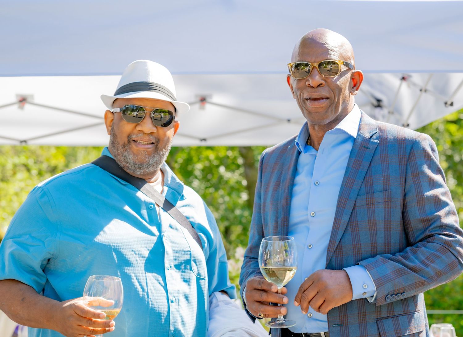 Two men holding wine glasses and smiling at an outdoor event under a white tent.