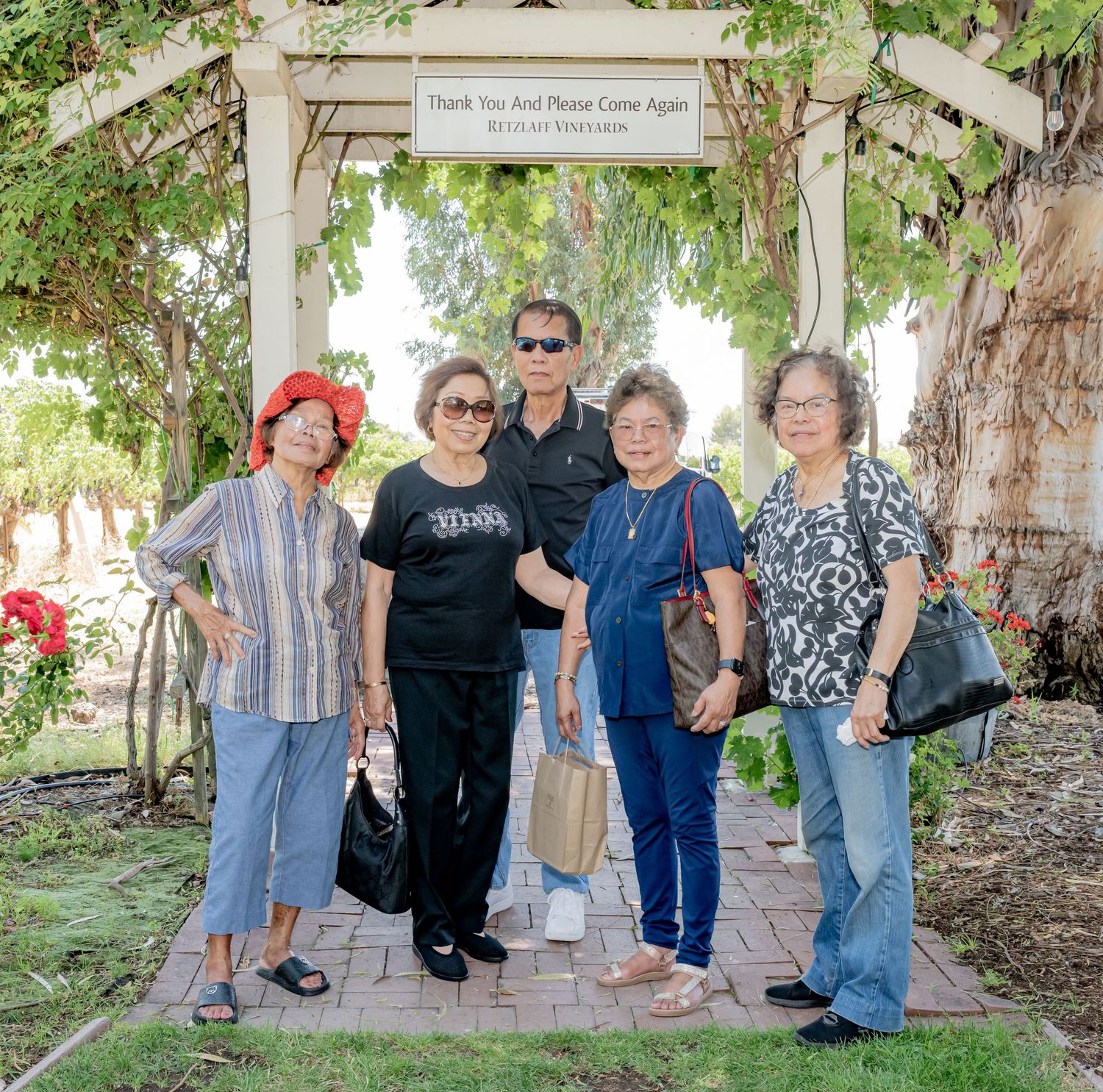 Five people pose under a garden arbor with a welcome sign at a scenic vineyard on a sunny day.