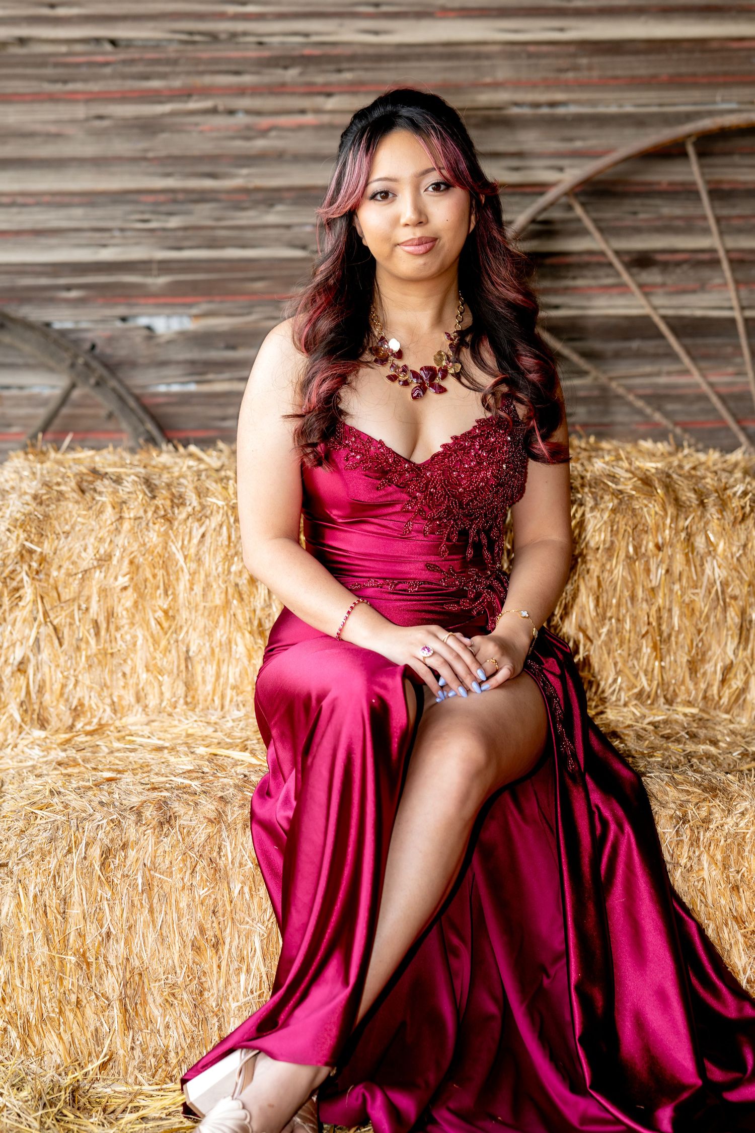 Beautiful woman in crimson satin formal gown poses on hay bales in rustic barn.