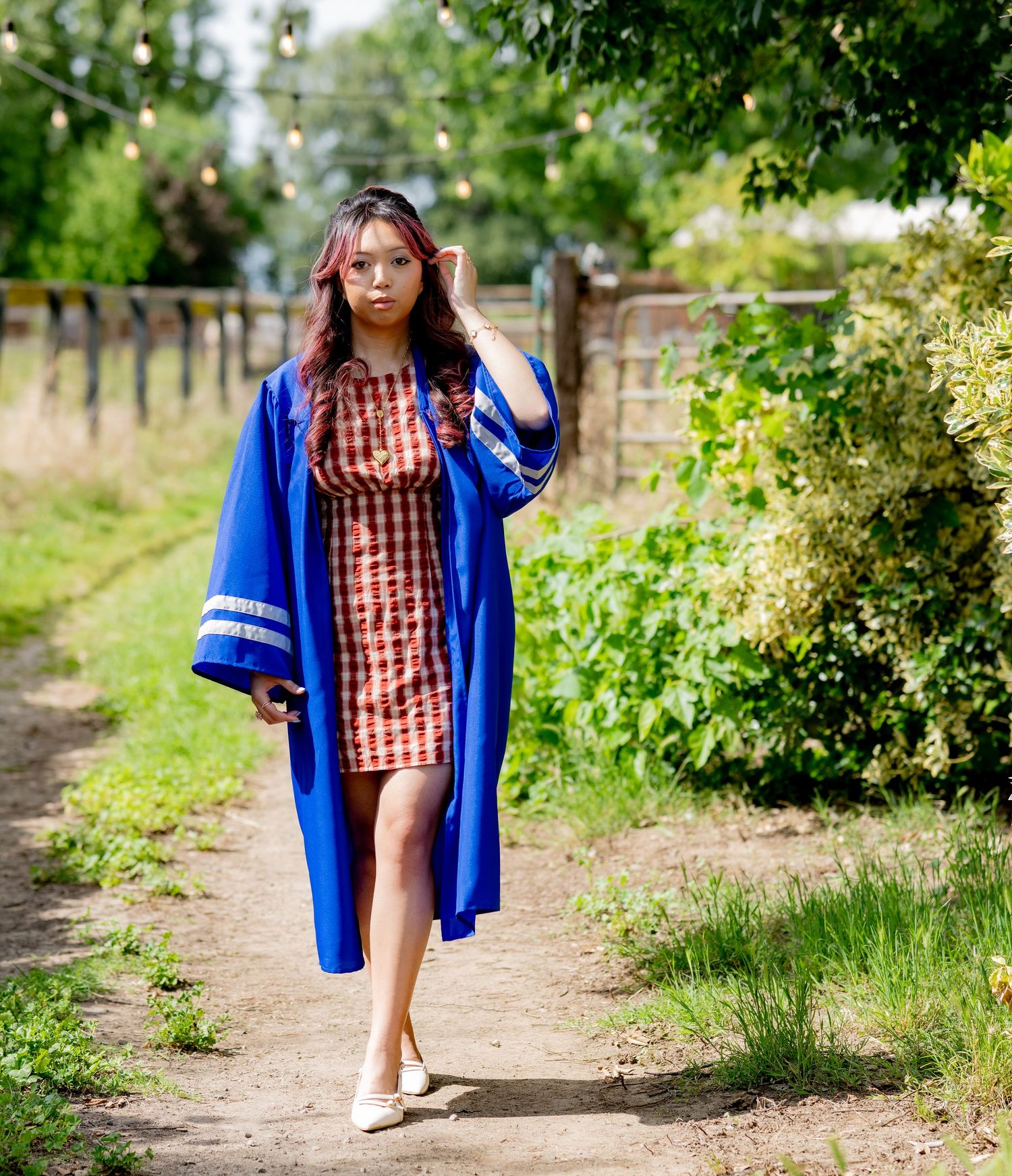 Graduate in royal blue commencement robe and red plaid dress poses on a scenic outdoor pathway.