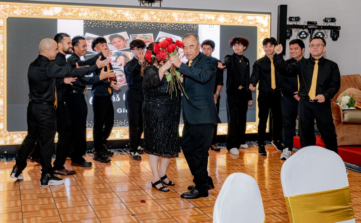 Older couple embraces with red roses surrounded by young men in black suits at birthday celebration.