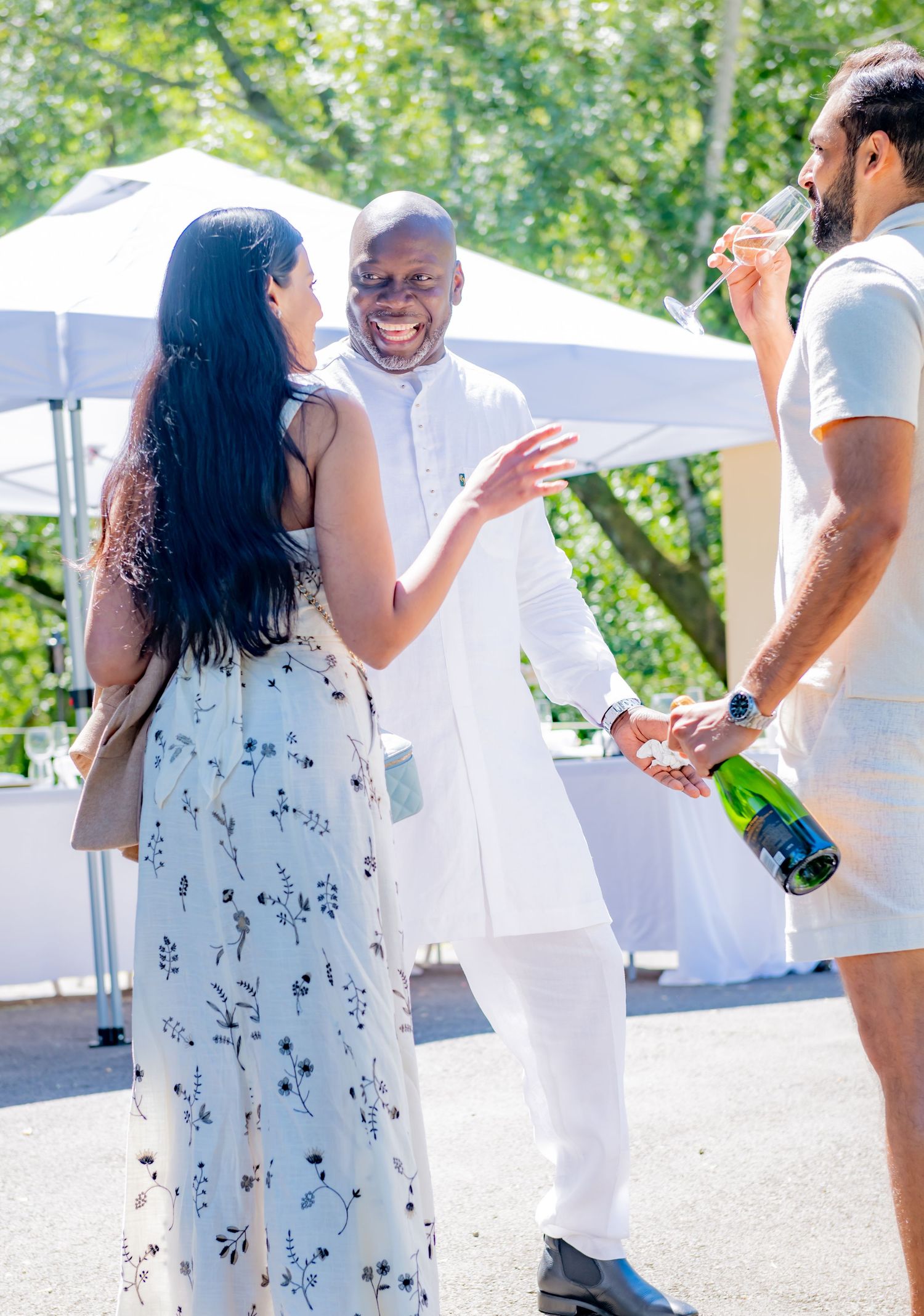 Three guests socializing outdoors at a sunny event, man in white pouring champagne.
