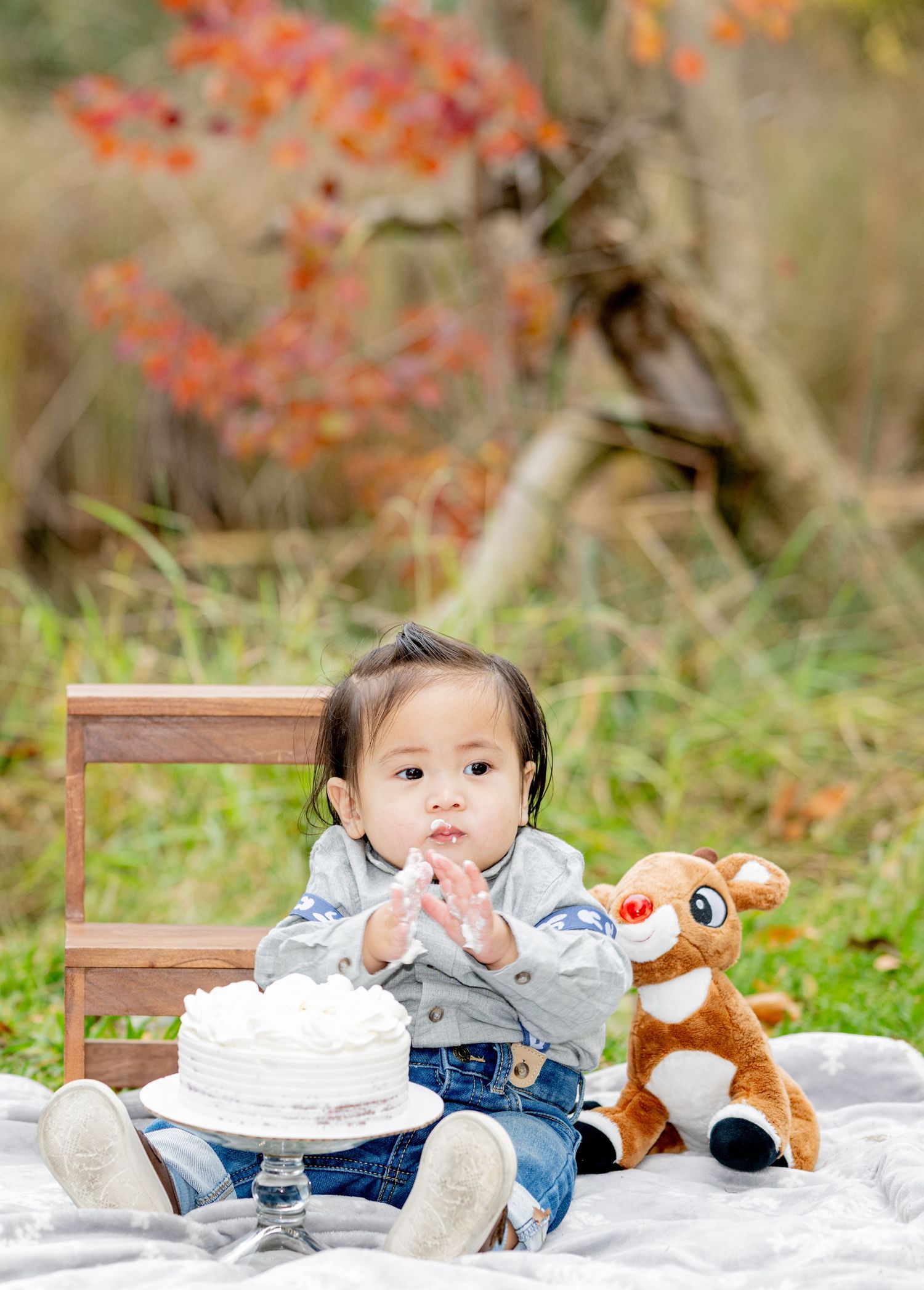 Baby boy in suspenders claps hands beside smash cake and stuffed reindeer toy outdoors.