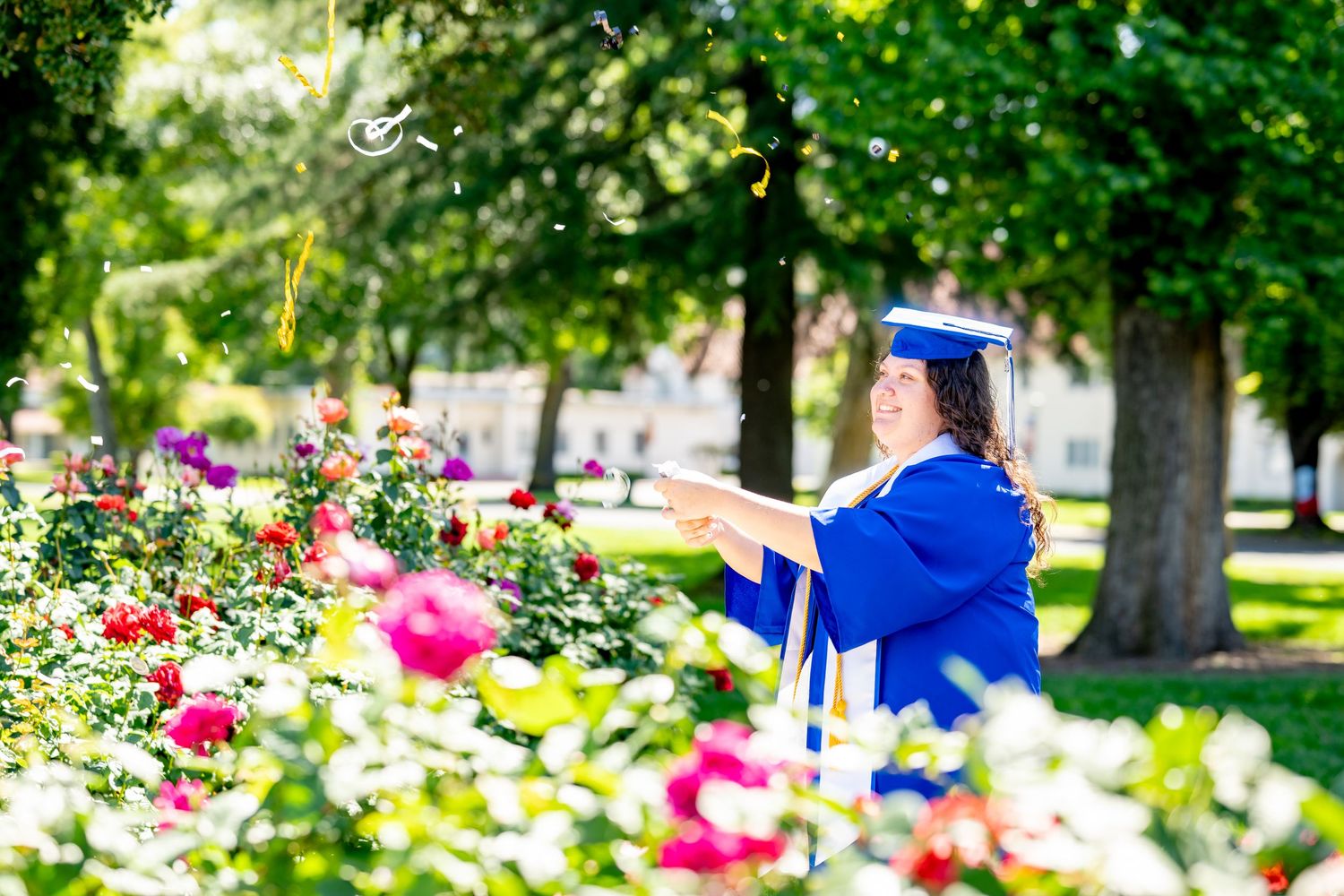 Female graduate in blue cap and gown tossing tassel among colorful rose garden outdoors.