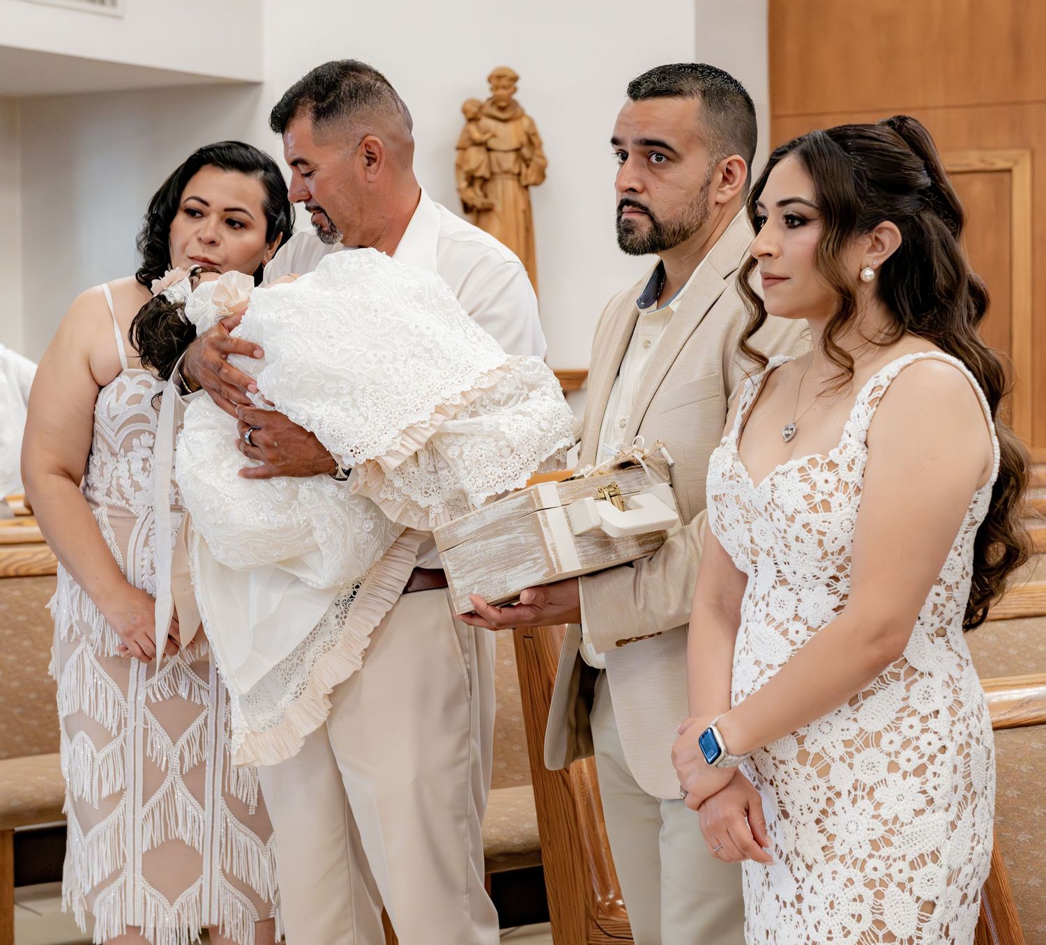 Family stands together holding baby in white christening gown and baptism gifts during a Catholic baptism ceremony.