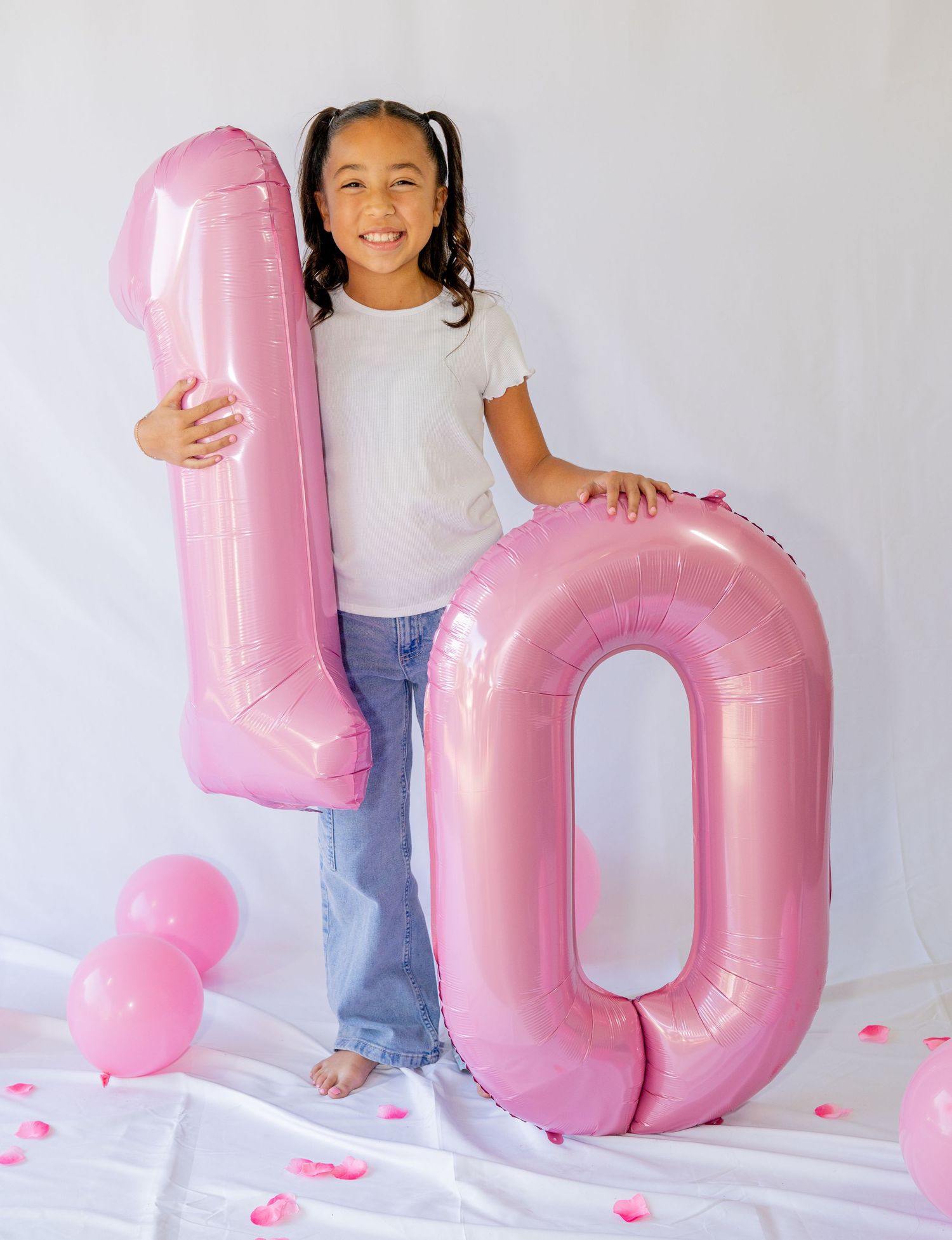 Smiling girl posing with large pink number 10 balloon for tenth birthday photo on white background.