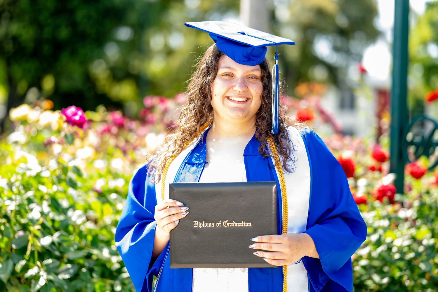Smiling graduate in blue regalia proudly displays her Diploma of Graduation outdoors.