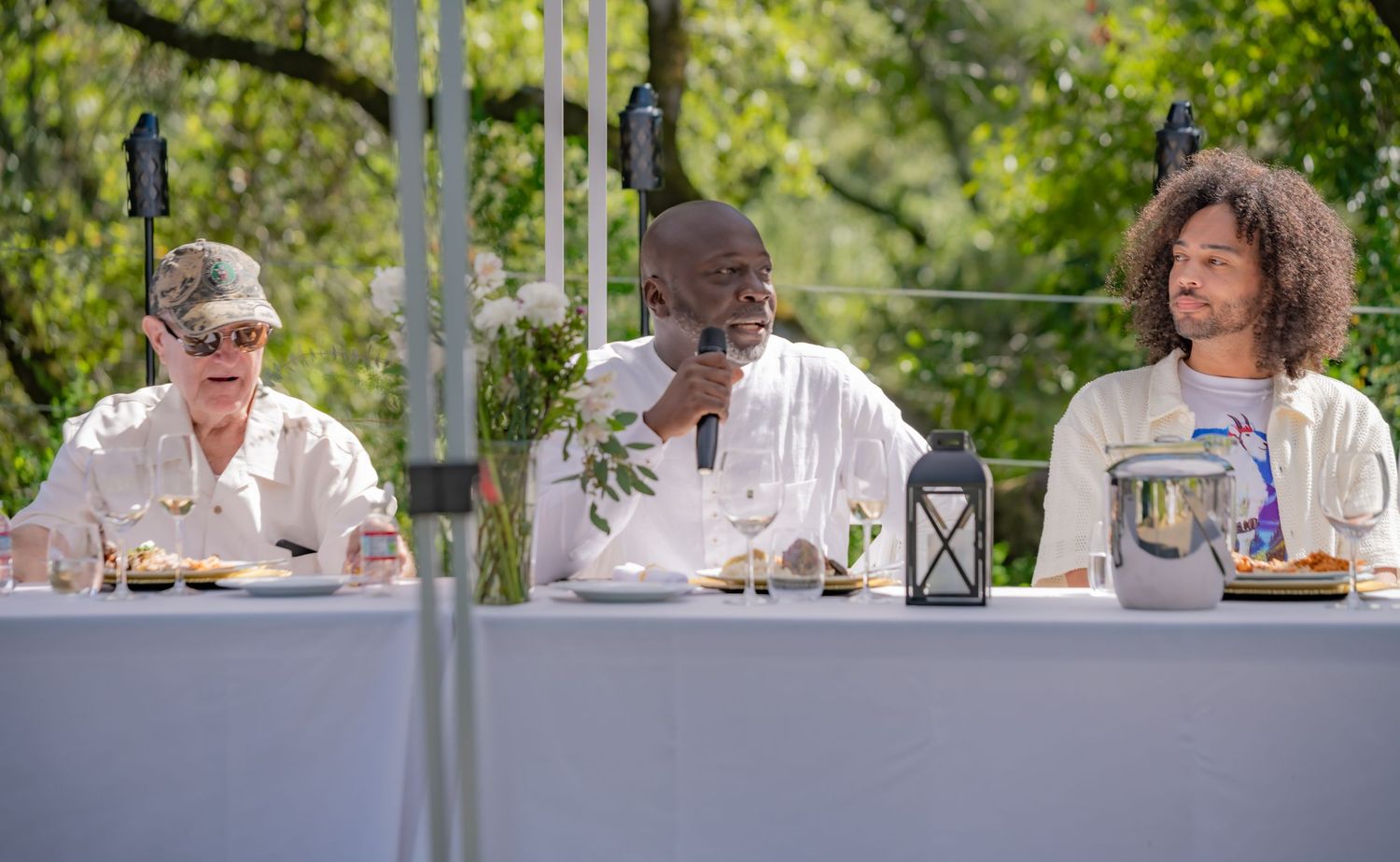 Three men seated at an outdoor banquet table, center man speaking into microphone.