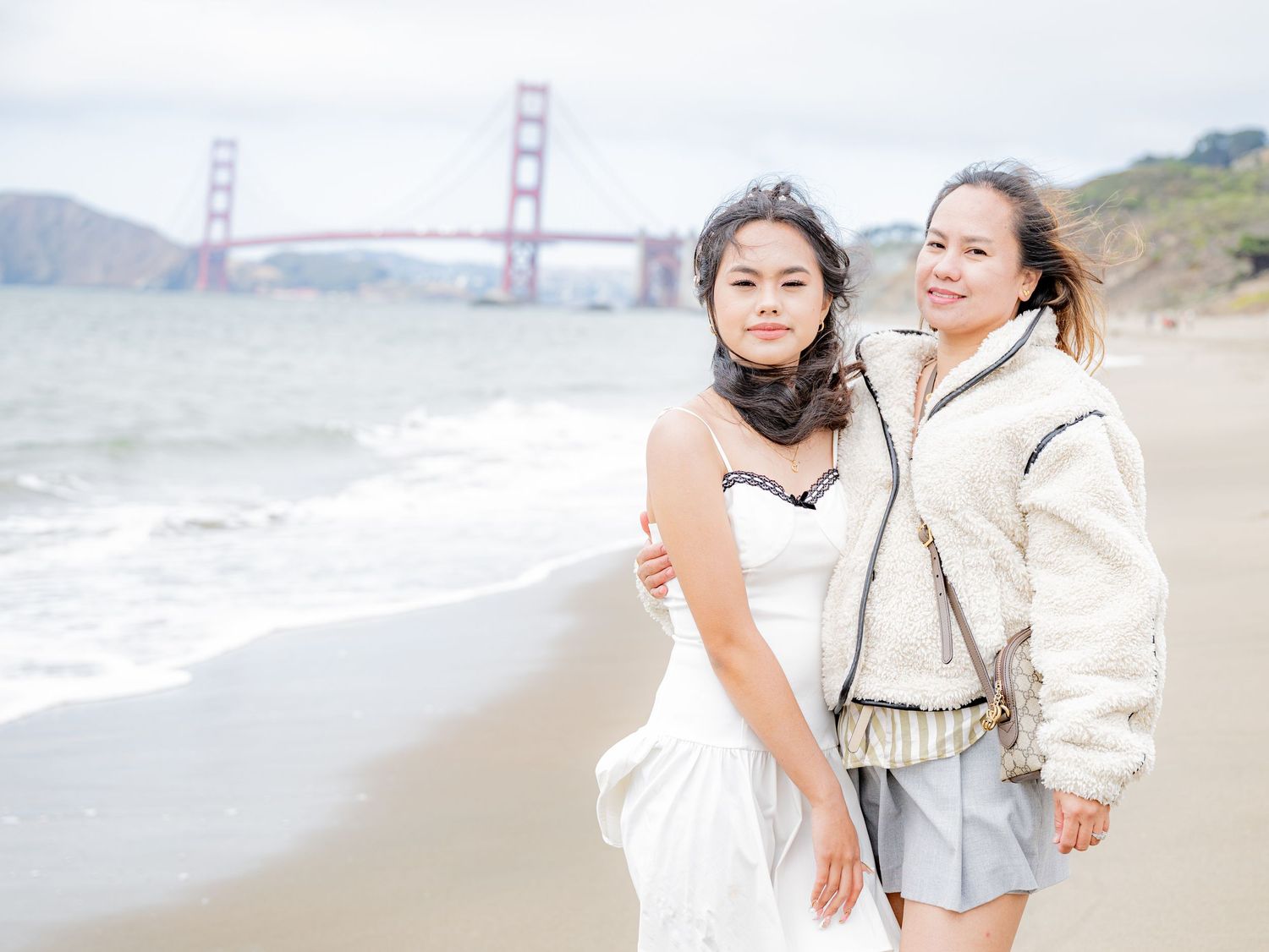 Two women pose together smiling on Baker Beach with Golden Gate Bridge in the background.