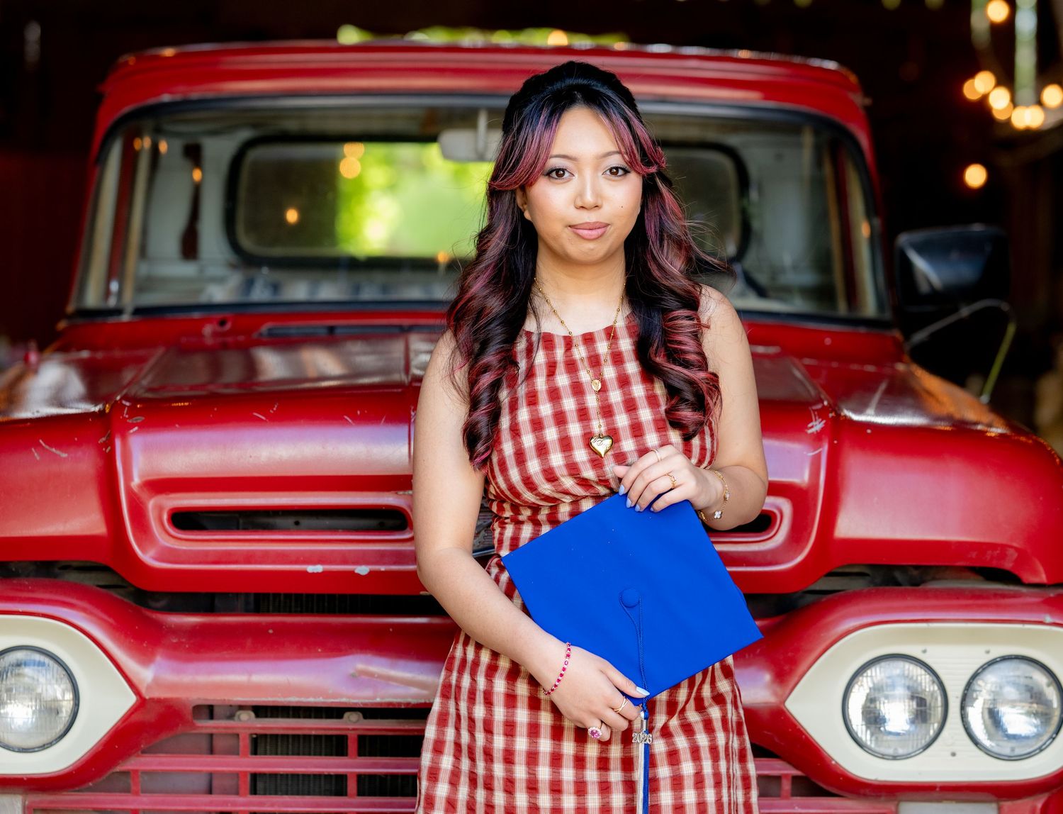Young woman in red plaid dress holding blue graduation cap in front of vintage red truck.