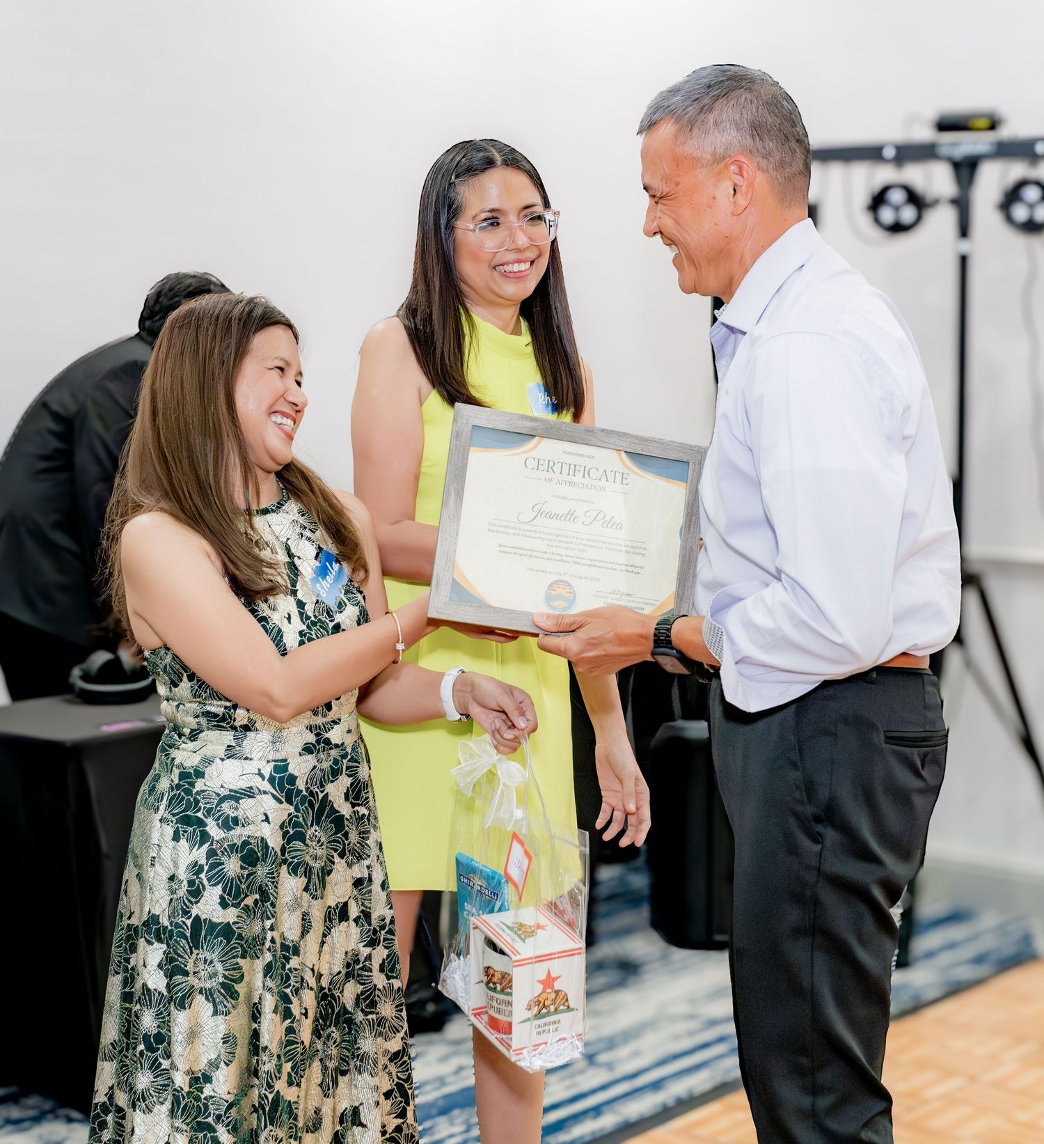 Two women smile while presenting a certificate to a man at a formal indoor recognition ceremony.
