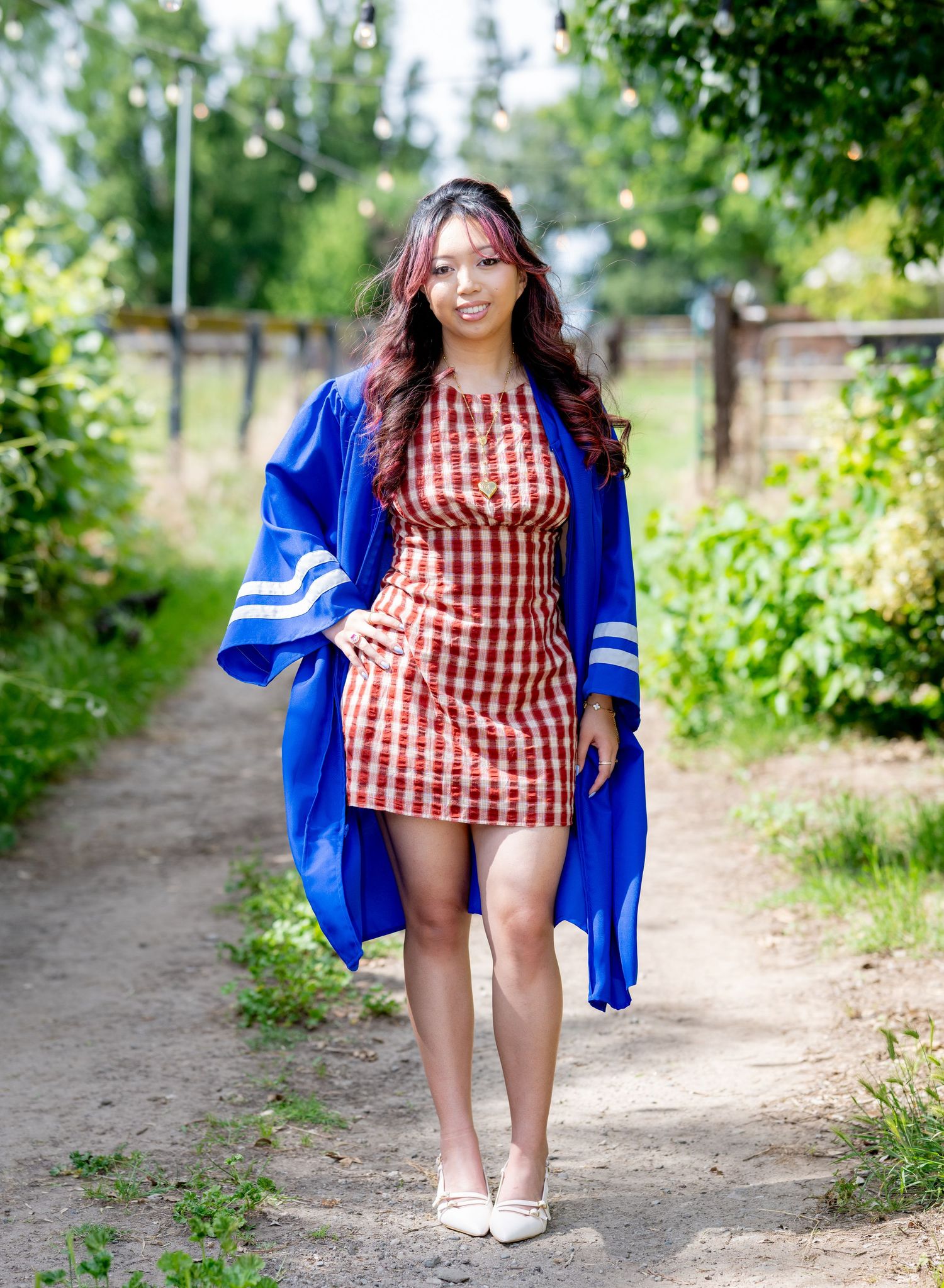 Graduate walking dirt path in blue gown over red gingham mini dress and white heels.