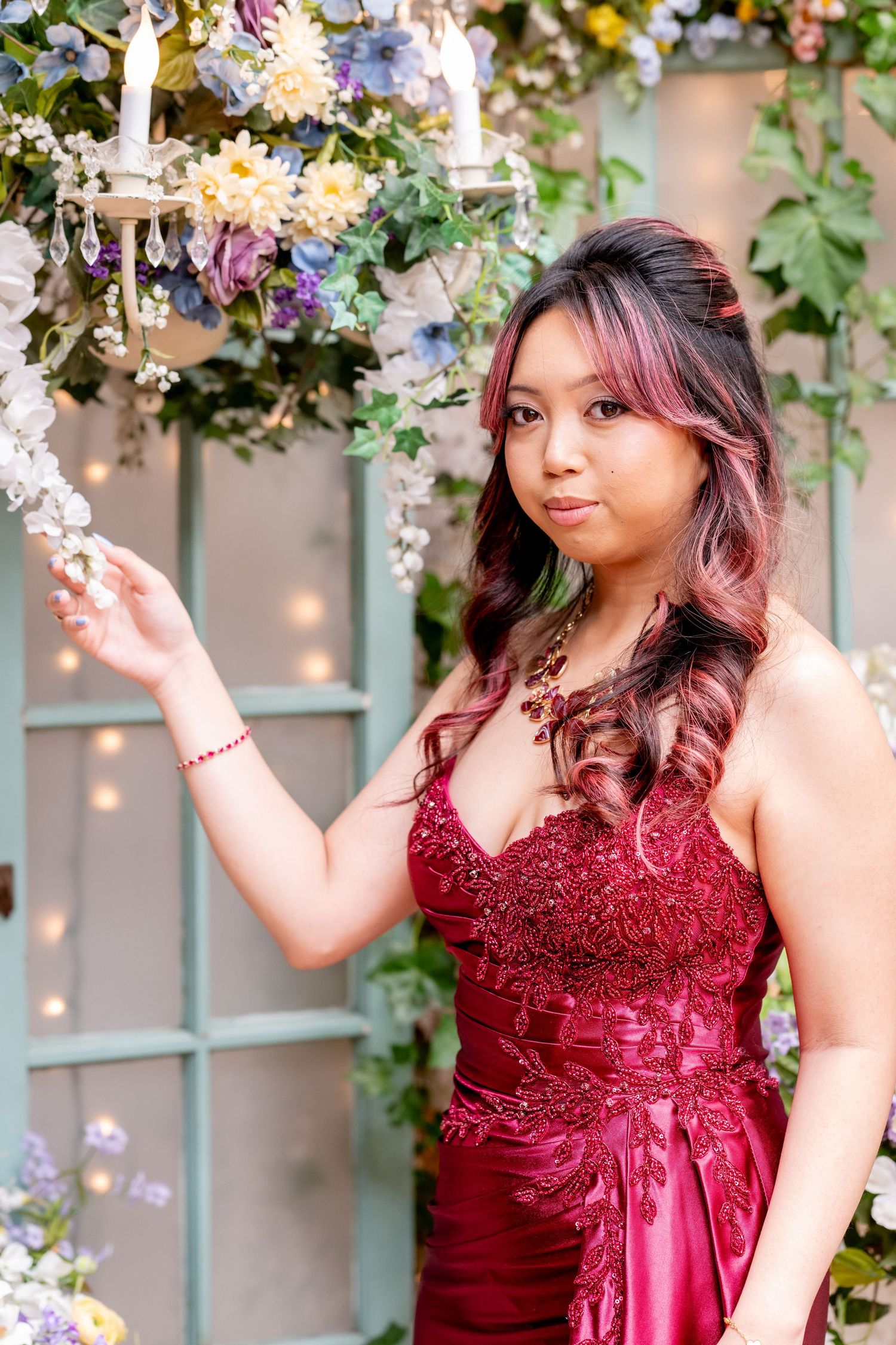 Woman in deep red lace-bodice formal gown poses beside chandelier with colorful floral arrangement backdrop.