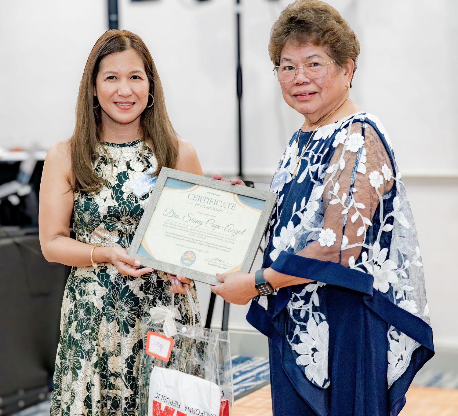 Two women smiling while holding a certificate of achievement at a formal recognition ceremony.