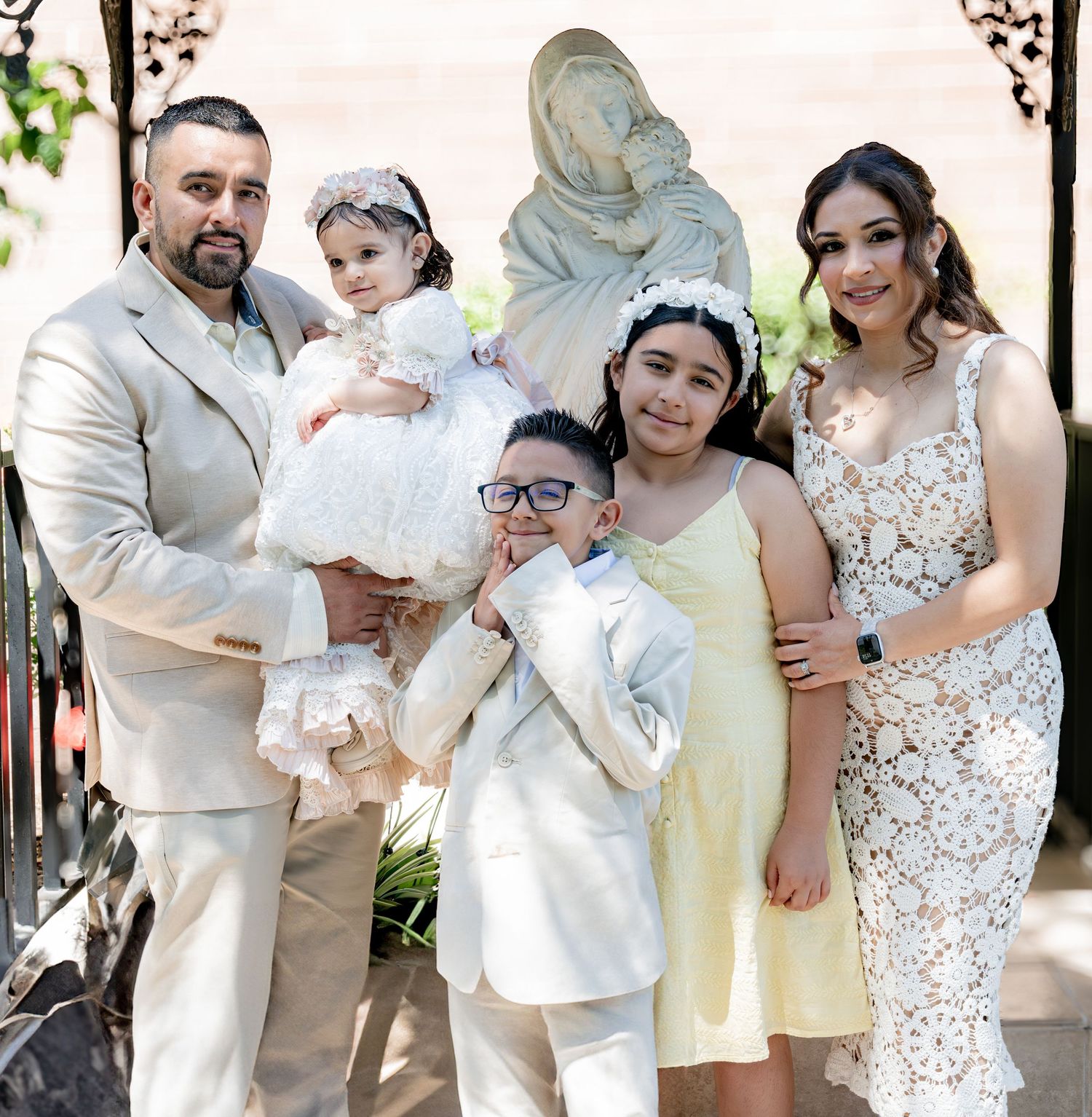 Family portrait with baby in baptism gown, two children, and parents by Virgin Mary statue.