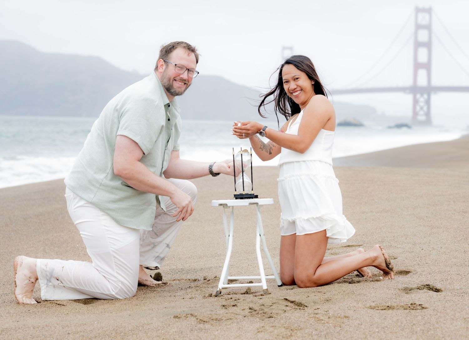 Man proposes to woman kneeling on Baker Beach with a small table and Golden Gate Bridge backdrop.